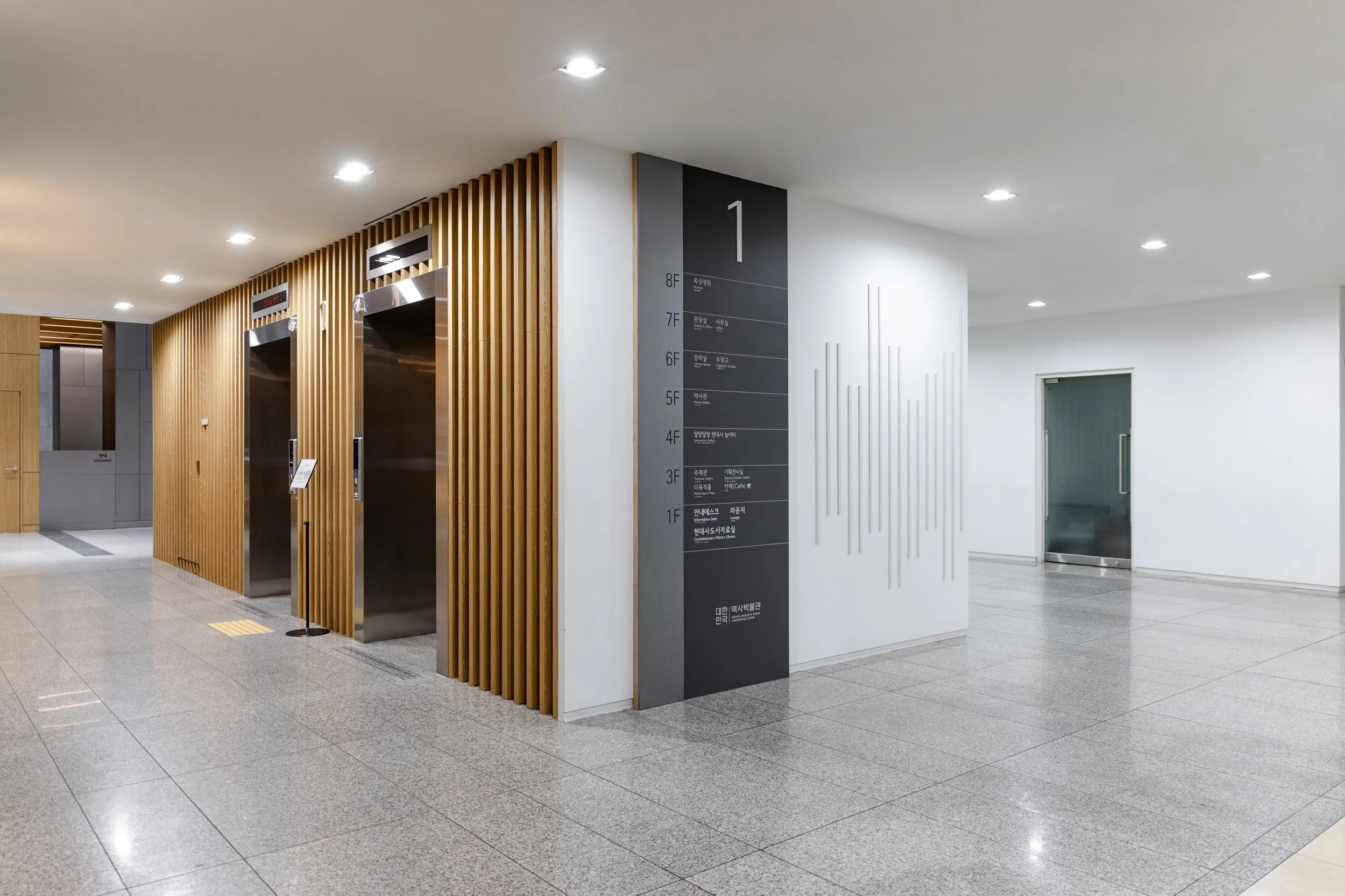 Interior of a modern building lobby with two elevators, wooden wall accents, gray flooring, and directory signs on the wall.