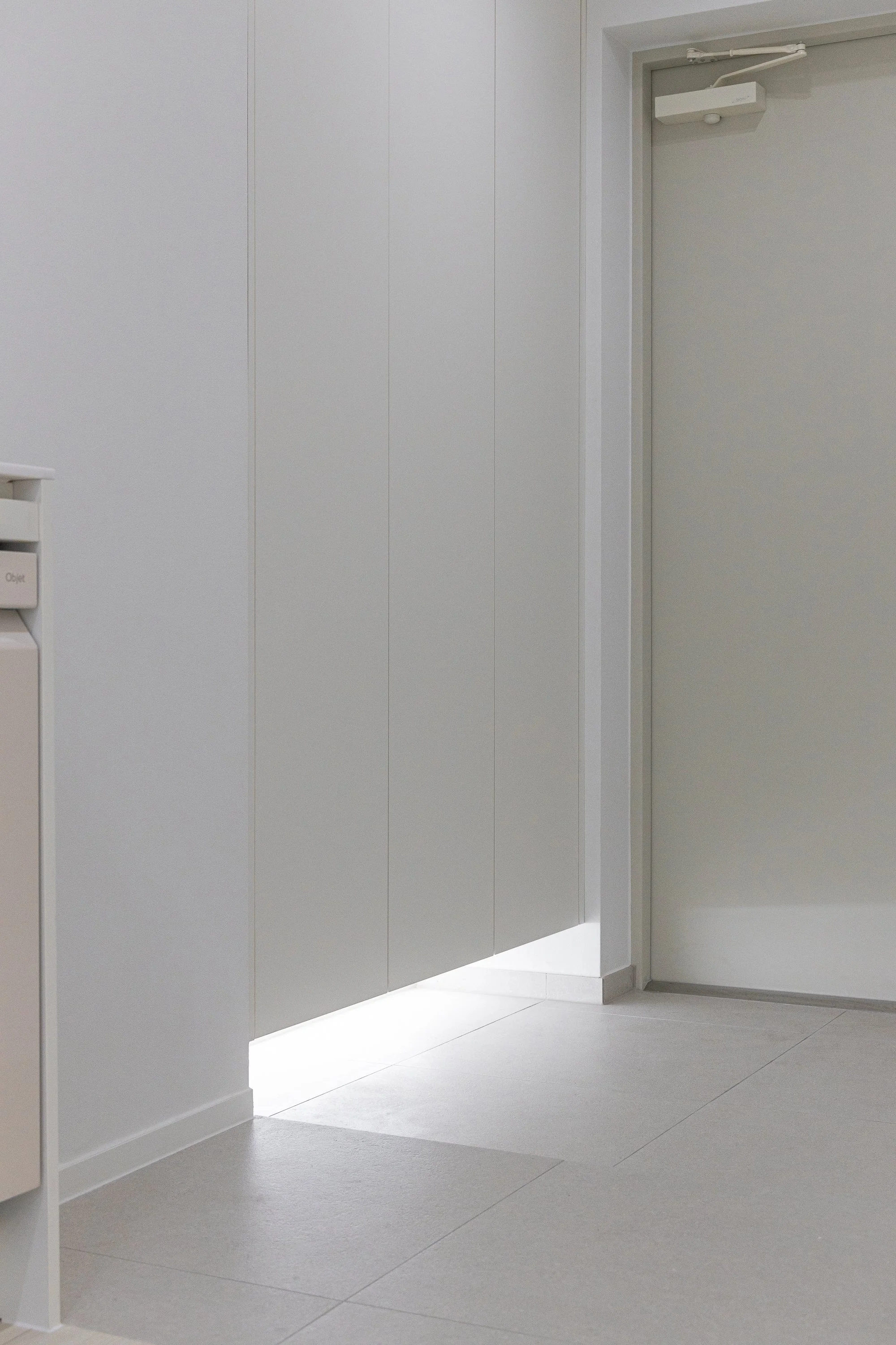 A white door with a metal door closer, closed inside a white room with minimal decor and gray flooring.