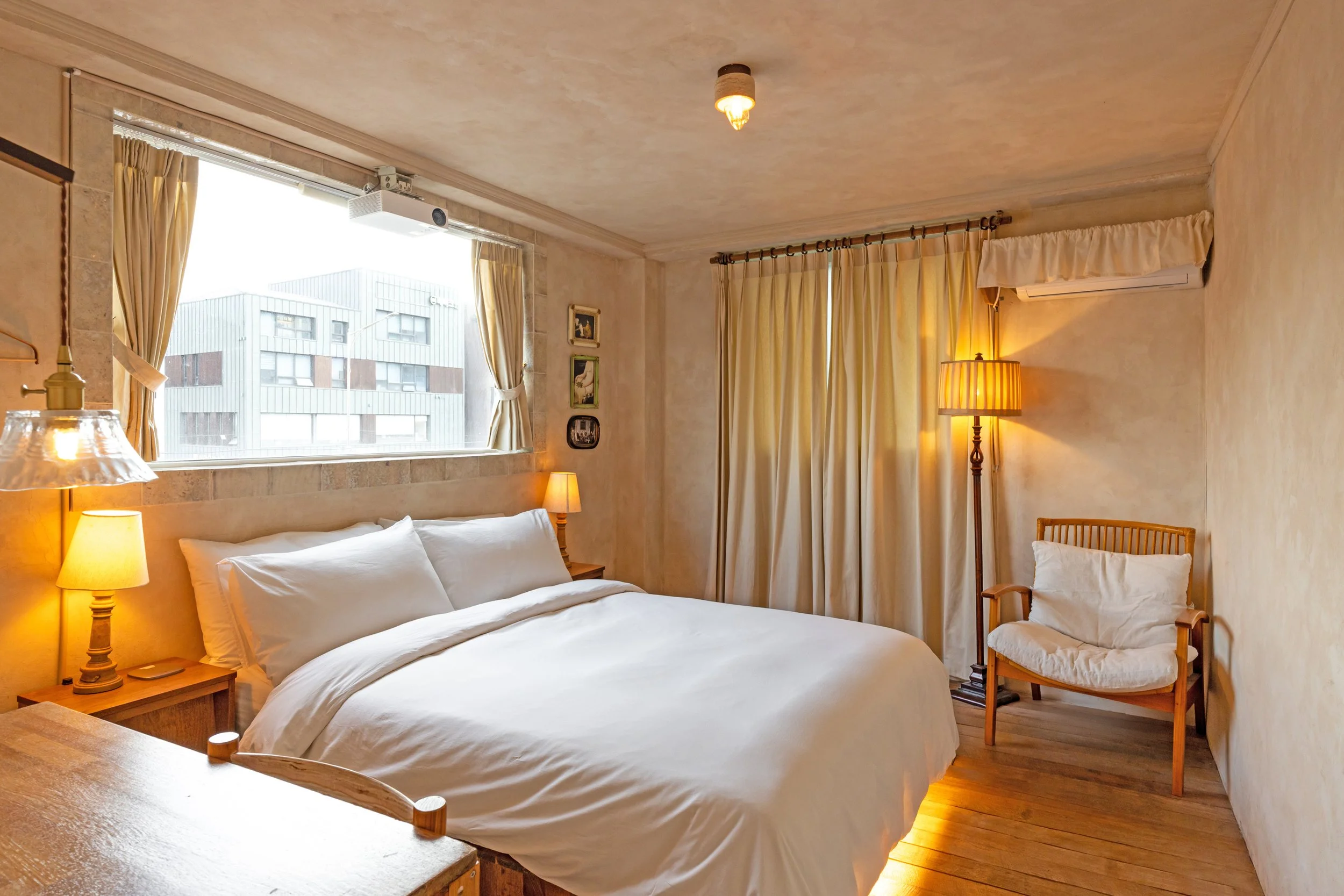Cozy bedroom with large bed, white bedding, wooden furniture, warm lighting, curtains, and a window showing a city building outside.
