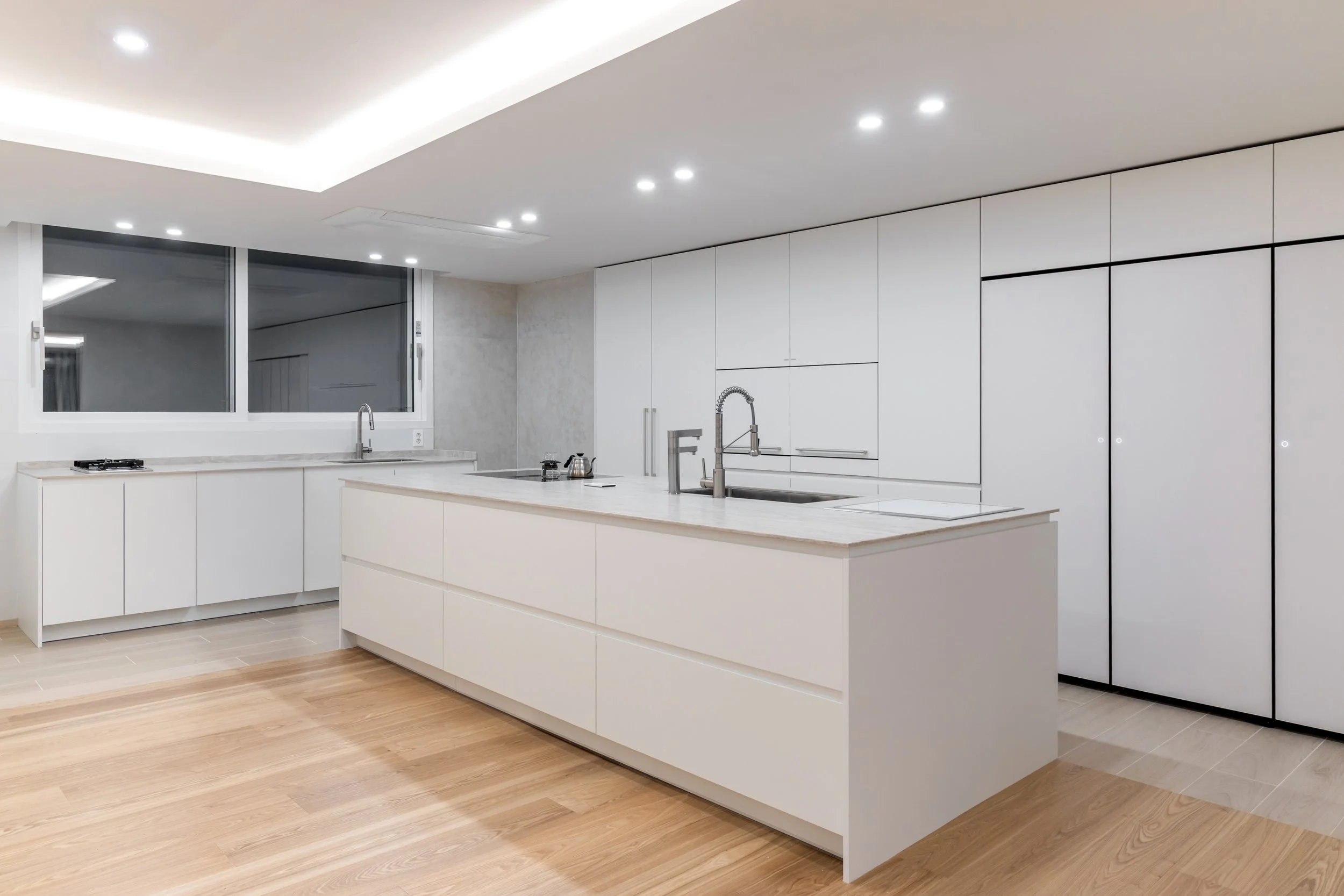 Modern kitchen with white cabinets, large island, marble countertops, stainless steel sink, and black-trimmed cabinets, illuminated by recessed ceiling lights.