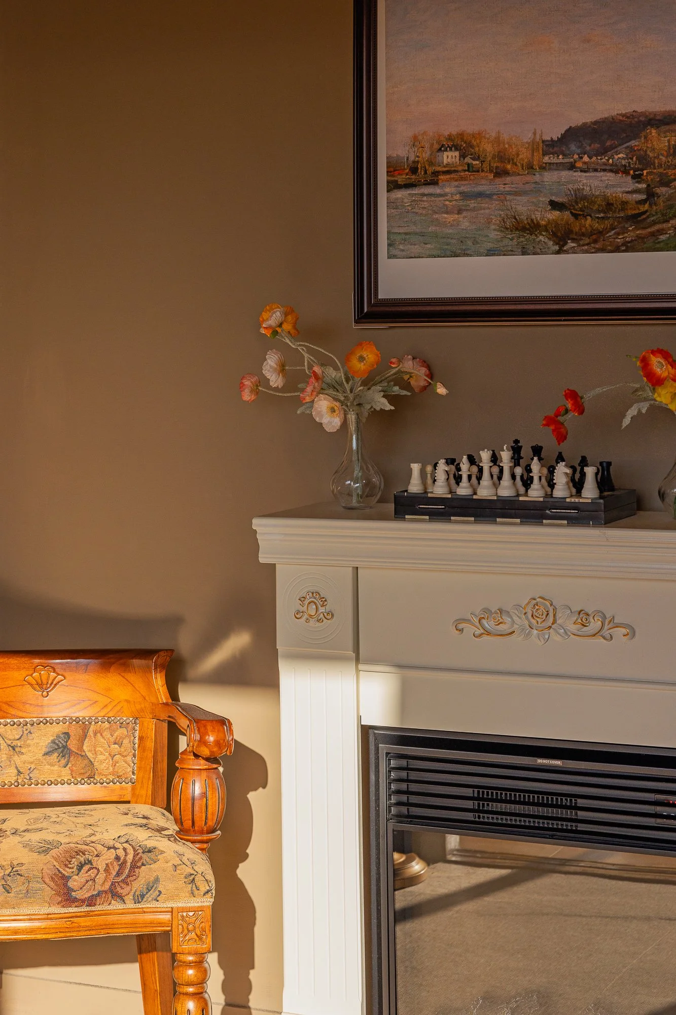 Close-up of a decorated fireplace mantel with a vase of flowers, a chess set, and a framed landscape painting above, next to a wooden chair with floral upholstery.
