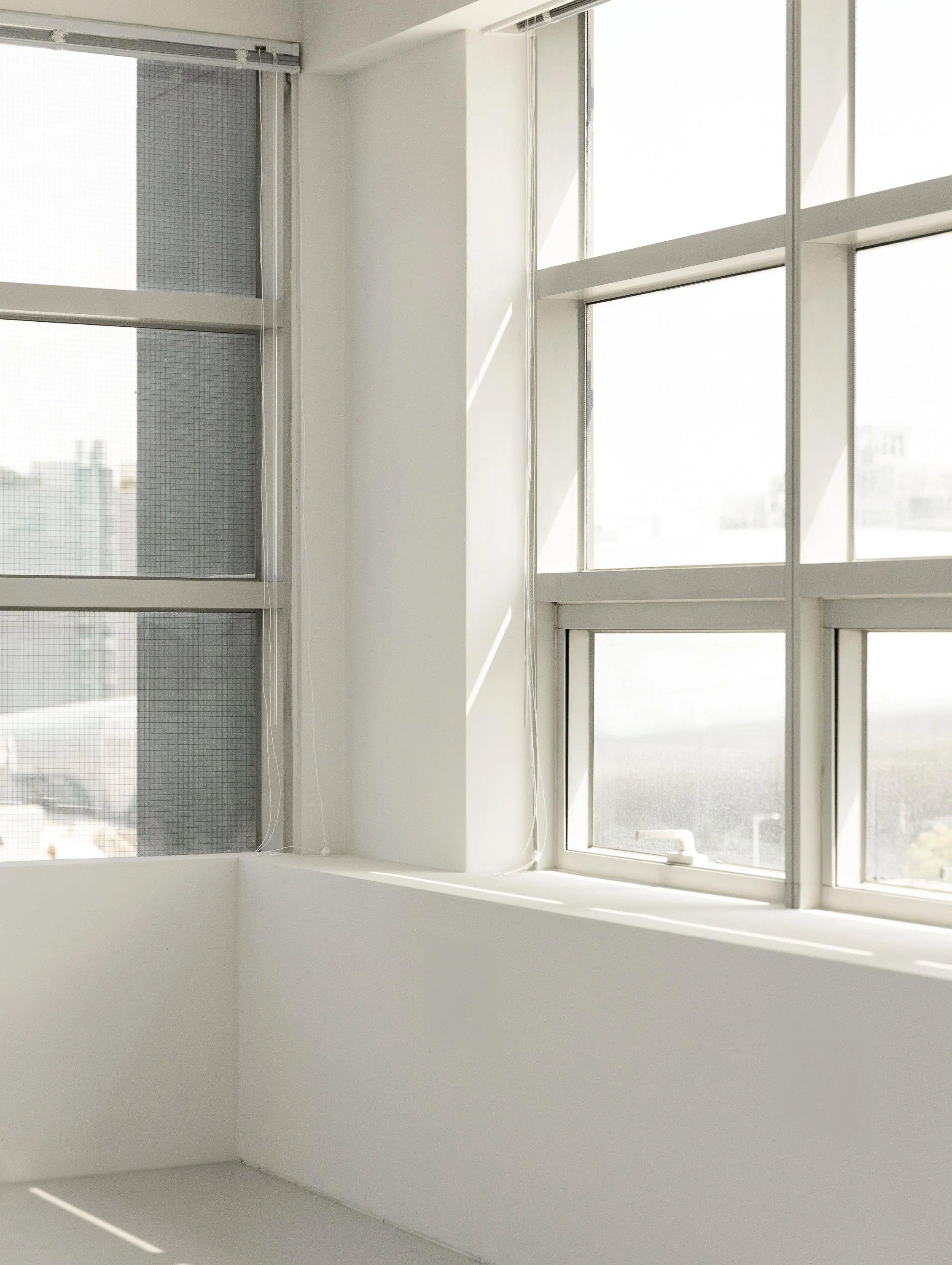 Sunlight coming through large office windows with white framing, revealing cityscape outside, with white walls and ledge inside.
