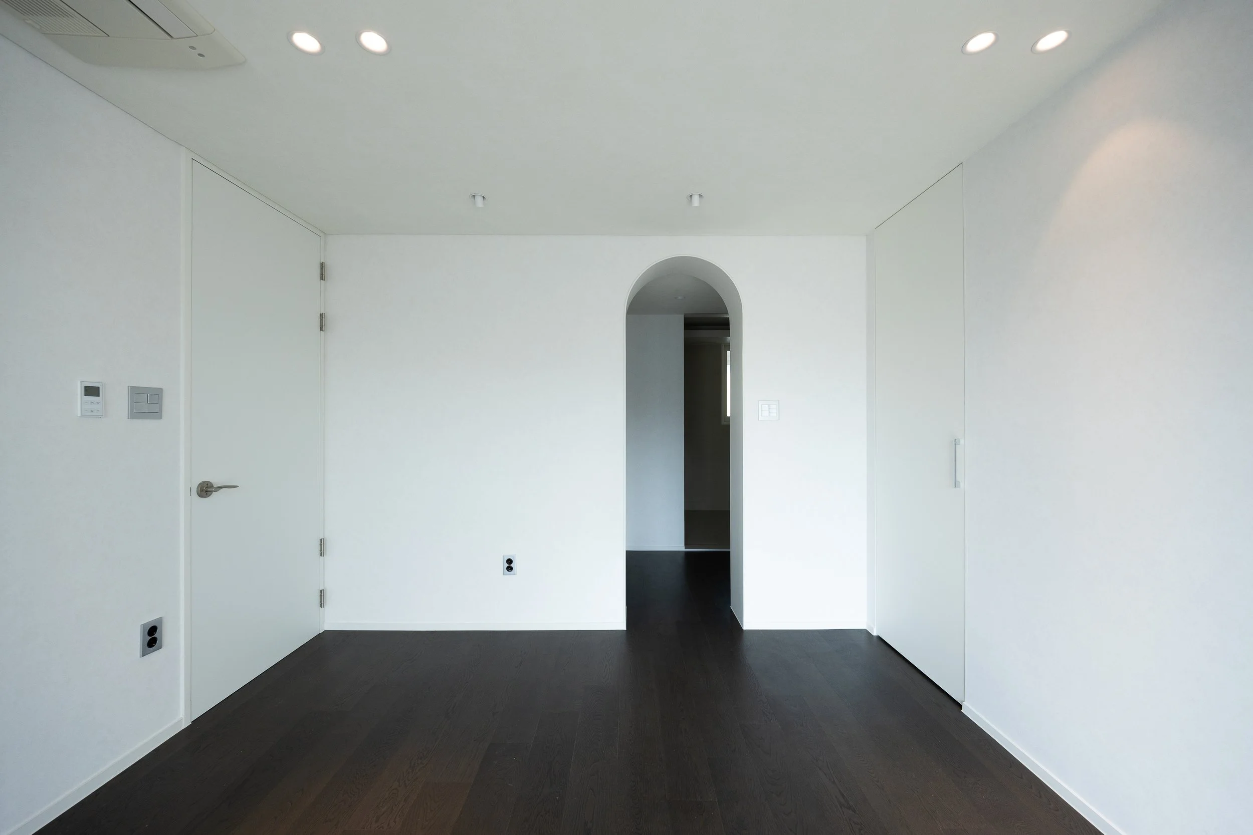 Empty modern room with white walls, dark hardwood floors, and an arched doorway leading to another space.
