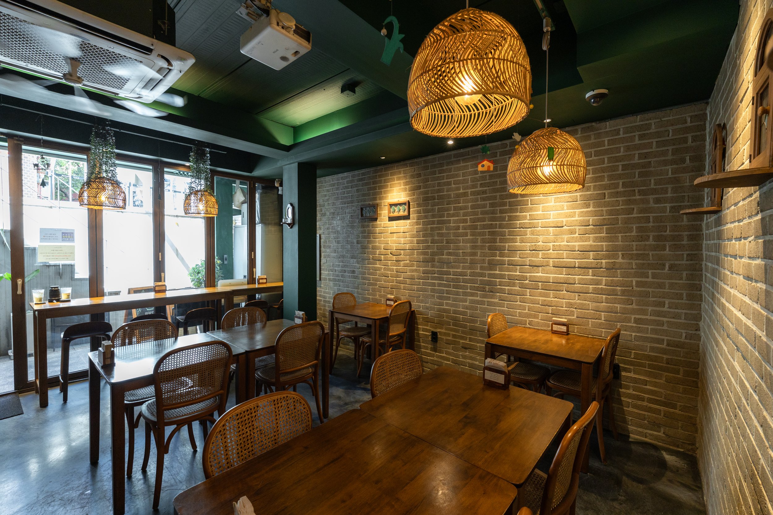 Interior of a cozy restaurant with wooden tables and rattan chairs, exposed brick walls, hanging wicker pendant lights, large windows, and small framed pictures on the wall.