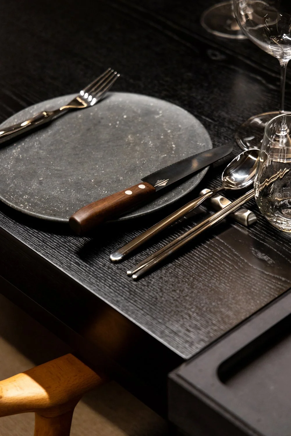 Empty black plate with a knife and spoon placed on a textured black placemat, set for a meal. Three glasses sit on the right side of the setup, on a black table.