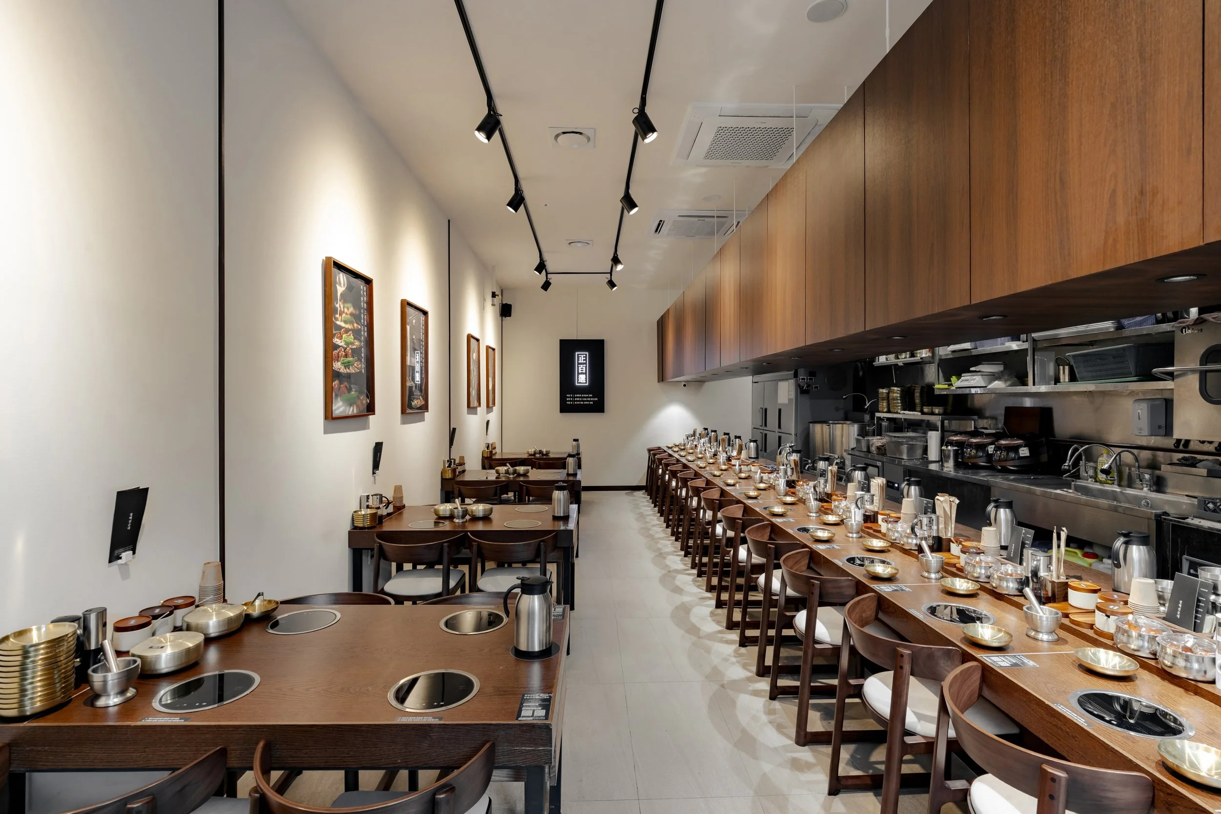 Interior of a modern Japanese restaurant with a long bar counter set with items, and several tables along the wall, decorated with framed food pictures and a digital menu display.
