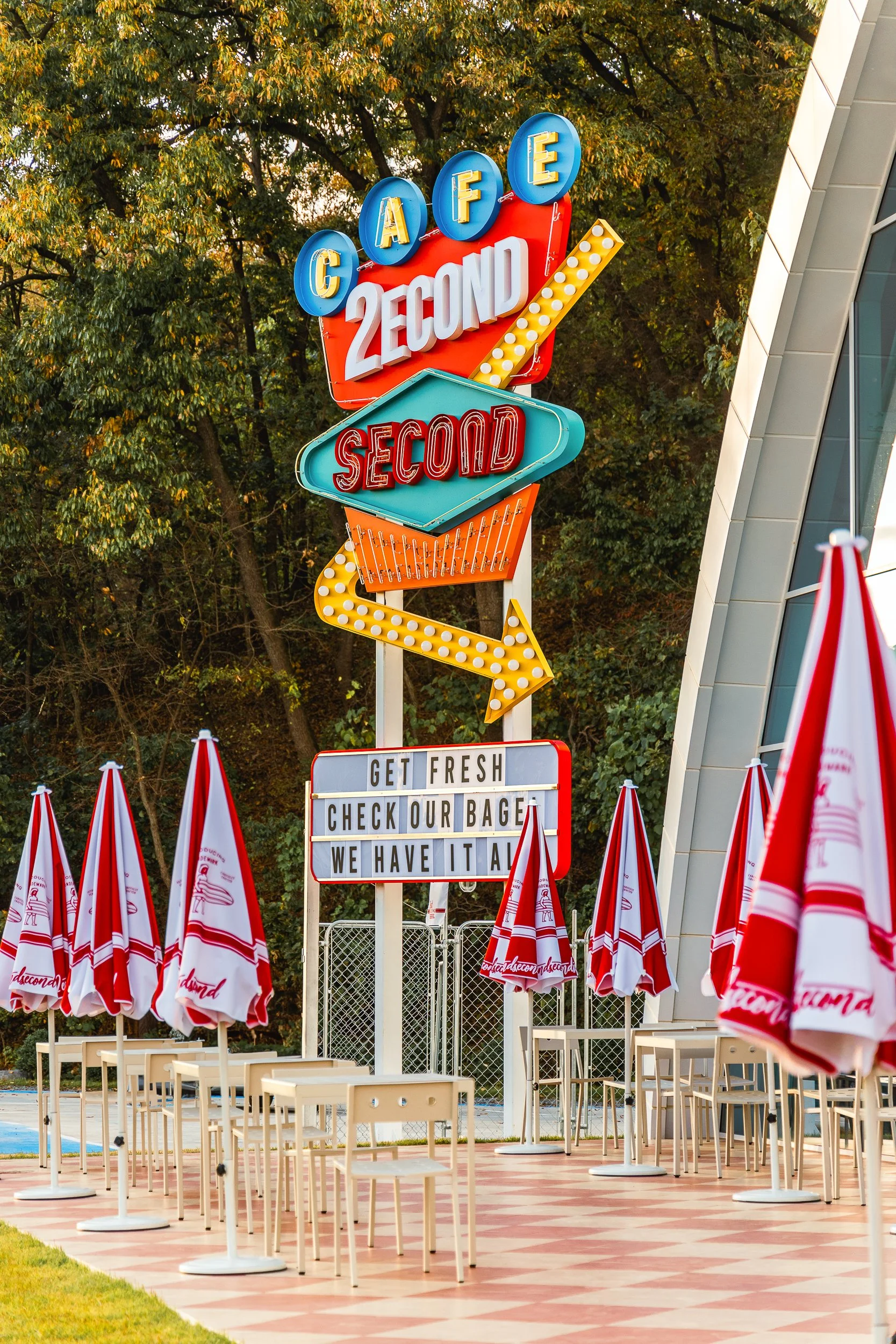 Colorful neon sign for 2nd Second, a dessert shop, with umbrellas and empty tables outside, and trees in the background.