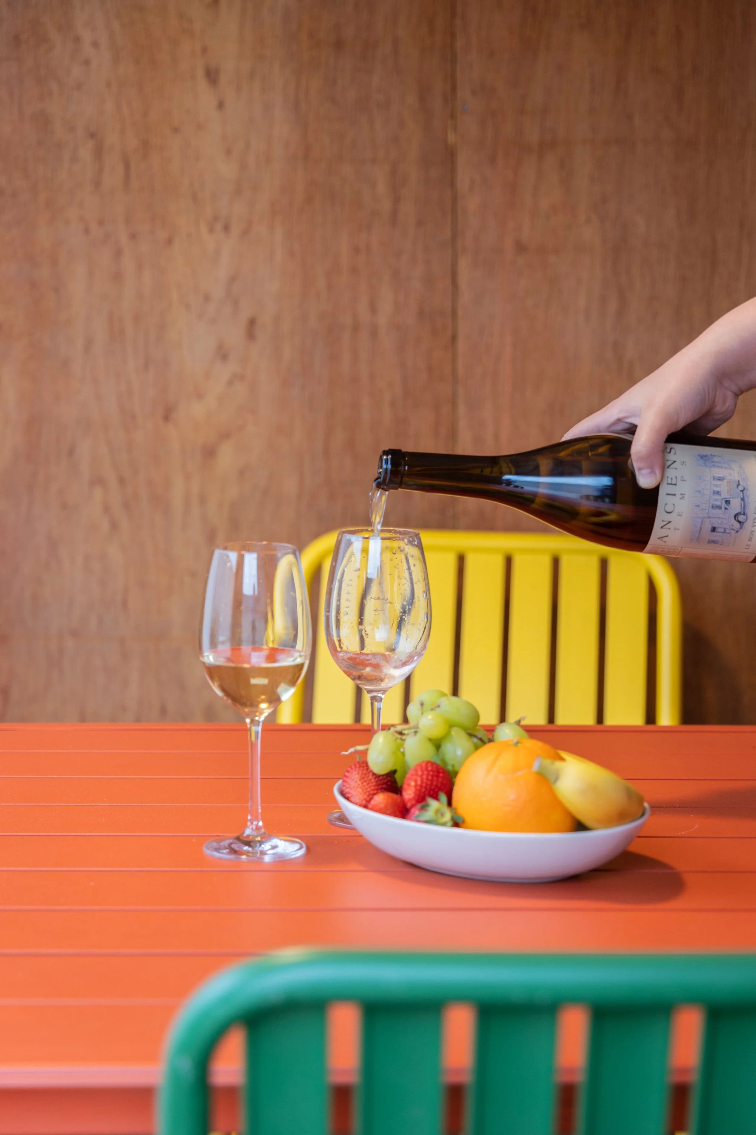 Person pouring wine into a glass on a table with a bowl of mixed fruit, including grapes, strawberries, an orange, and a banana, in the background.
