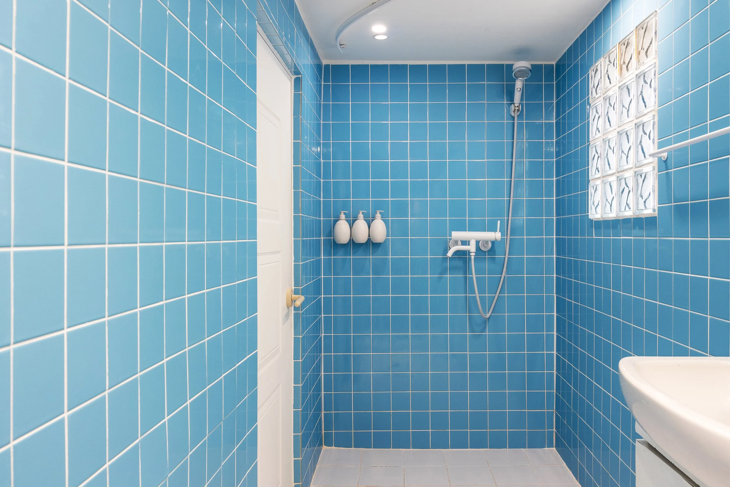 A bathroom with blue tiled walls, a white door, a shower head and controls, three white bottles on a shelf, a small window with glass blocks, and a white sink.