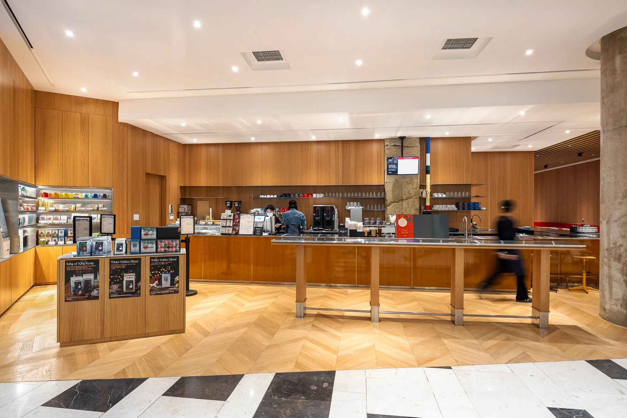 A modern coffee shop with a wooden interior, espresso machines, menu displays, a counter with snacks, and two staff members working behind the counter.