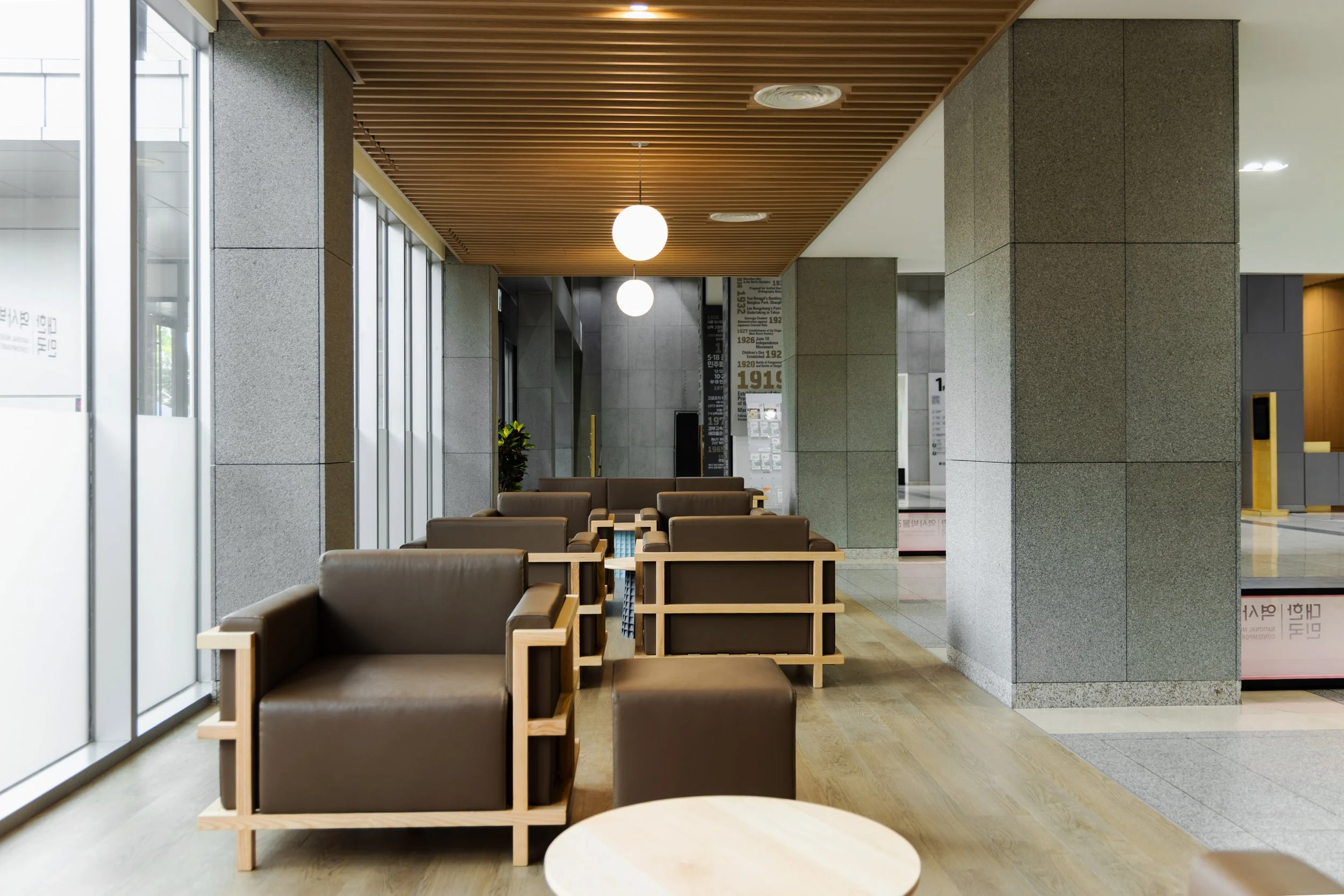 Empty modern lobby with brown leather armchairs, small round wooden table, large windows, wooden ceiling, stone pillars, and hanging spherical light fixtures.
