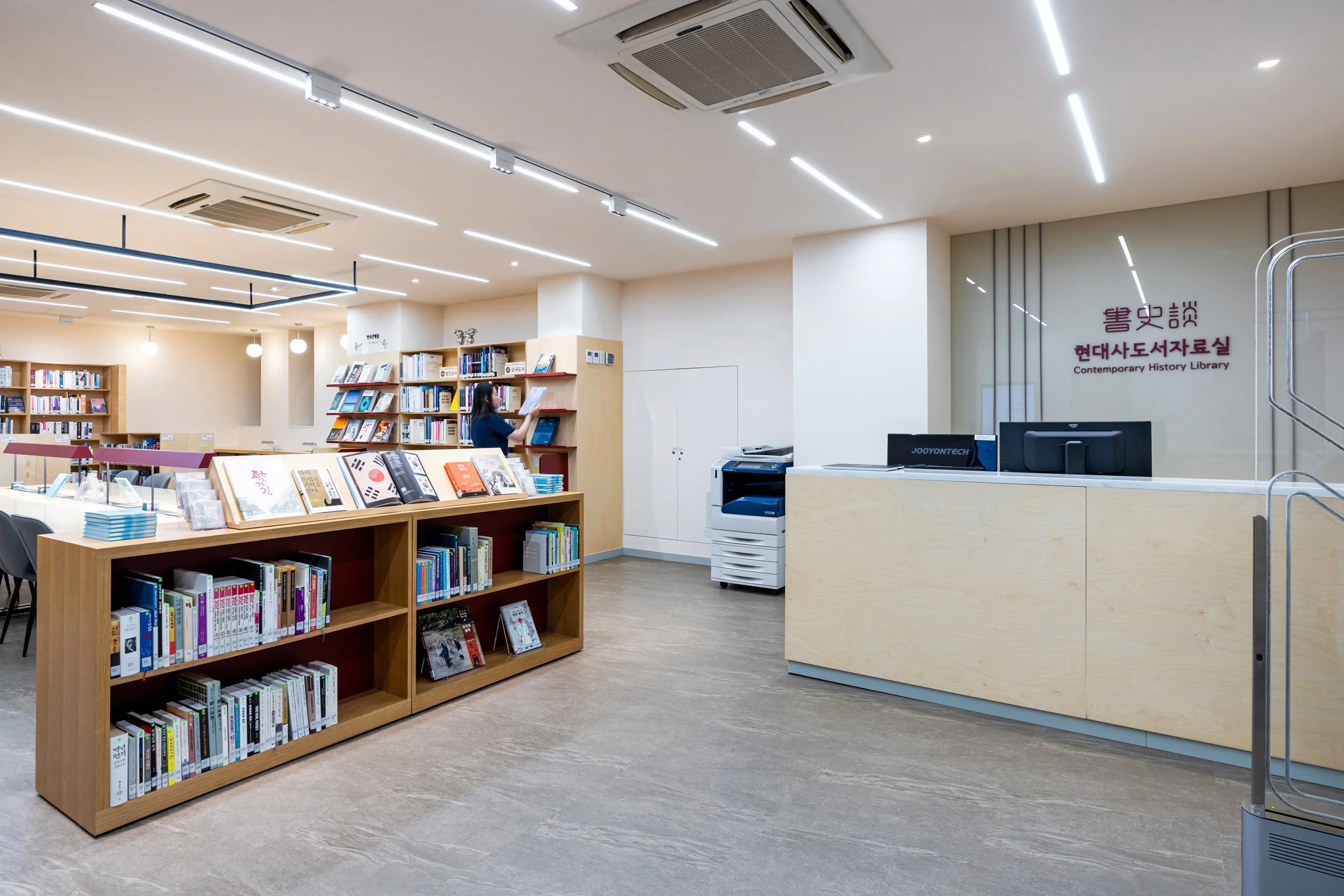 Interior of a modern library with wooden bookshelves, a person reading a book, a reception desk with computers, and a sign that reads 'Contemporary History Library' in both Korean and English.