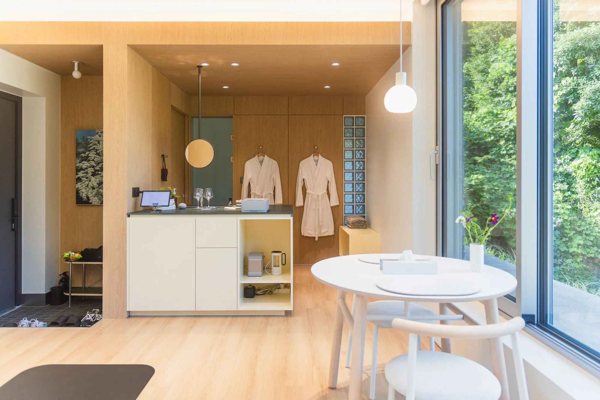 Modern kitchen and dining area with large window overlooking greenery, white dining table with a small flower arrangement, kitchen counter with robes hanging on the wall, and various kitchen appliances.