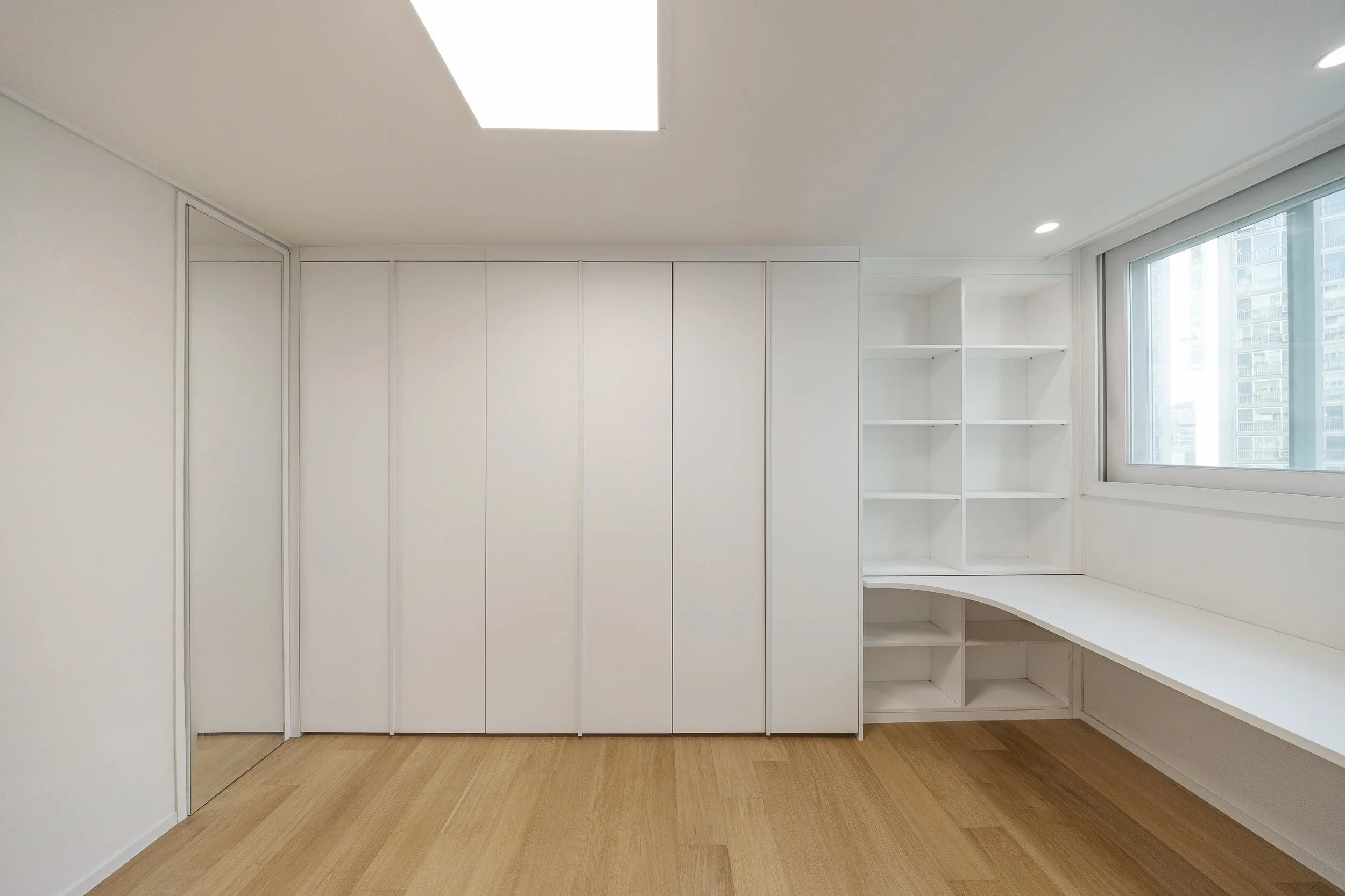 Empty room with white built-in shelves, a white desk by the window, wooden floor, and a skylight.