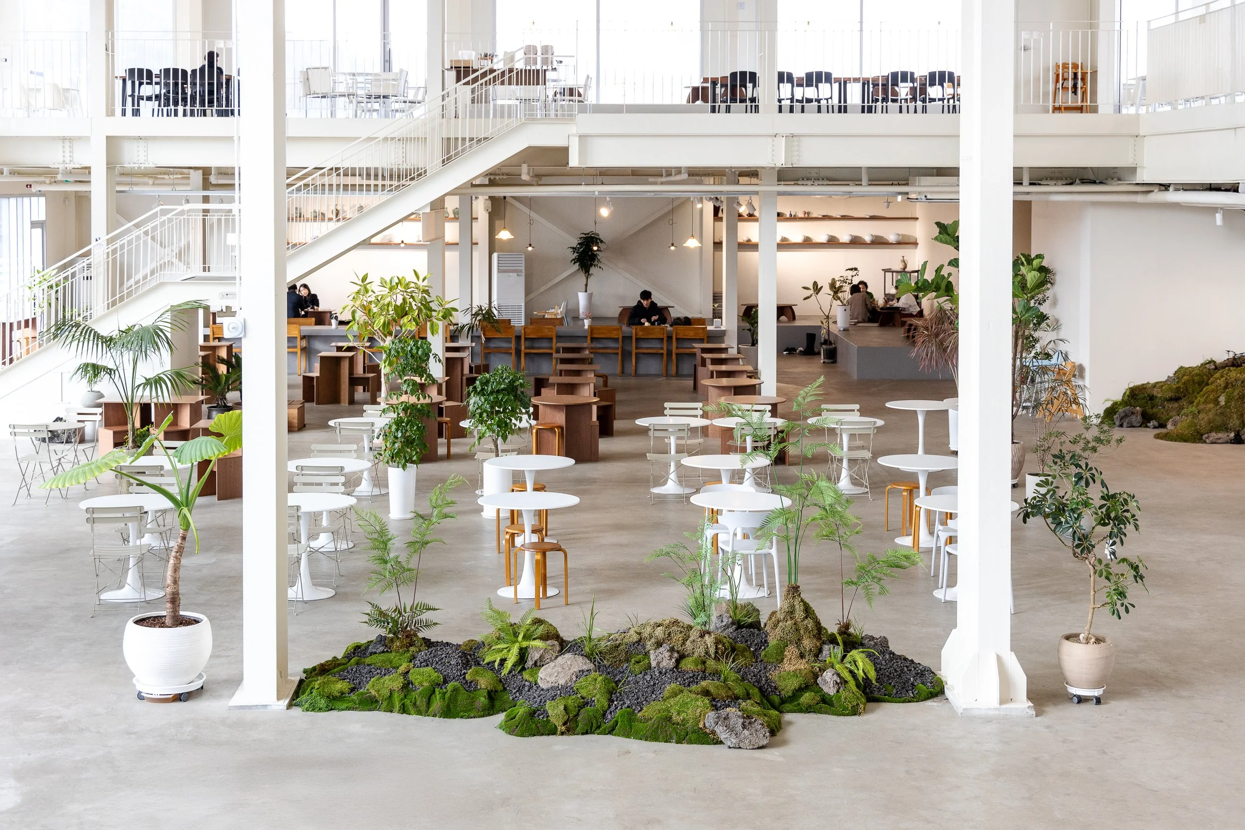 A spacious modern indoor cafe or co-working space with white tables and chairs, potted plants, and indoor greenery, featuring an open second floor with stairs, and people working or relaxing.