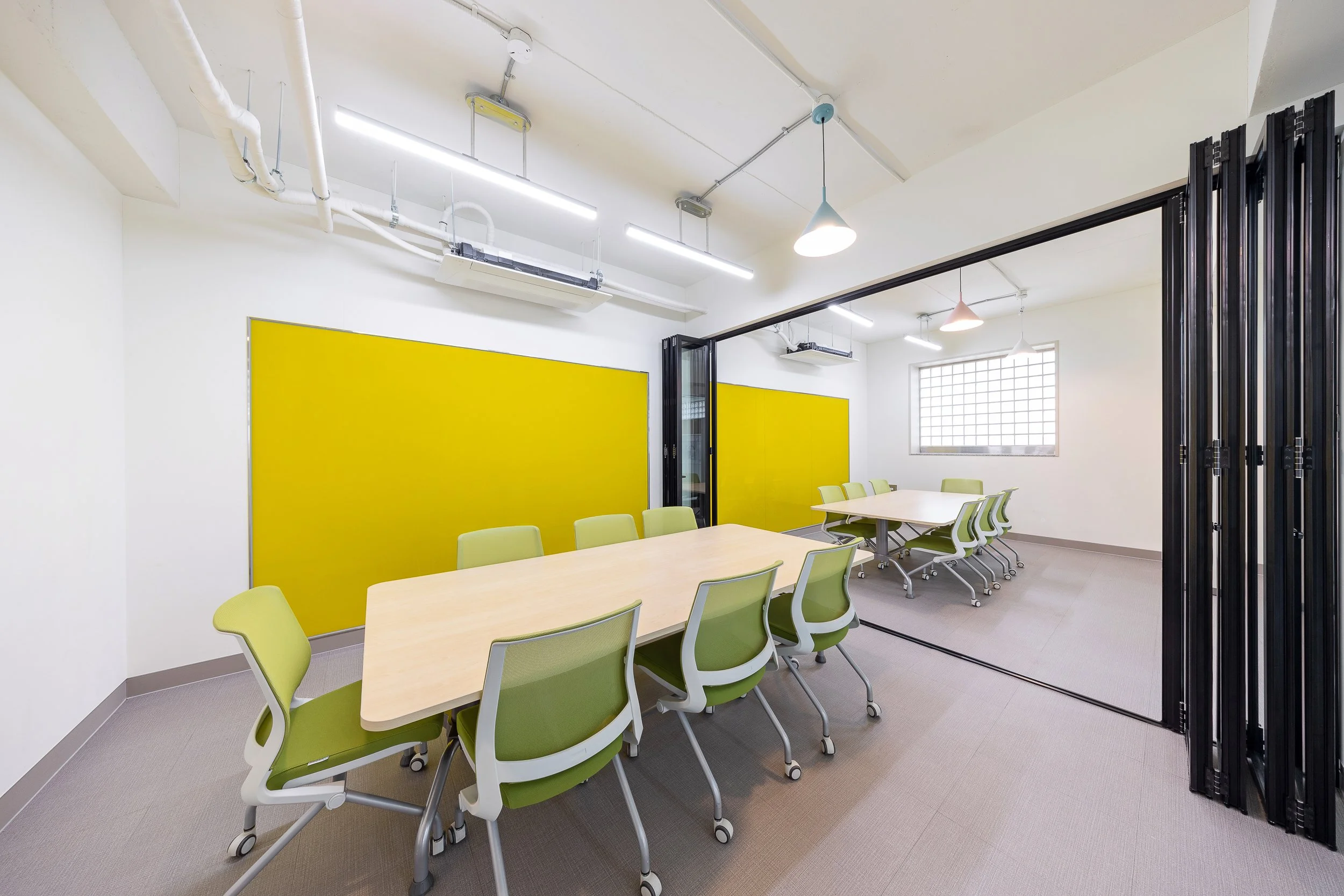 Conference room with two rectangular tables, green chairs, yellow wall panels, glass partition, white walls, window, ceiling lights, and exposed piping.