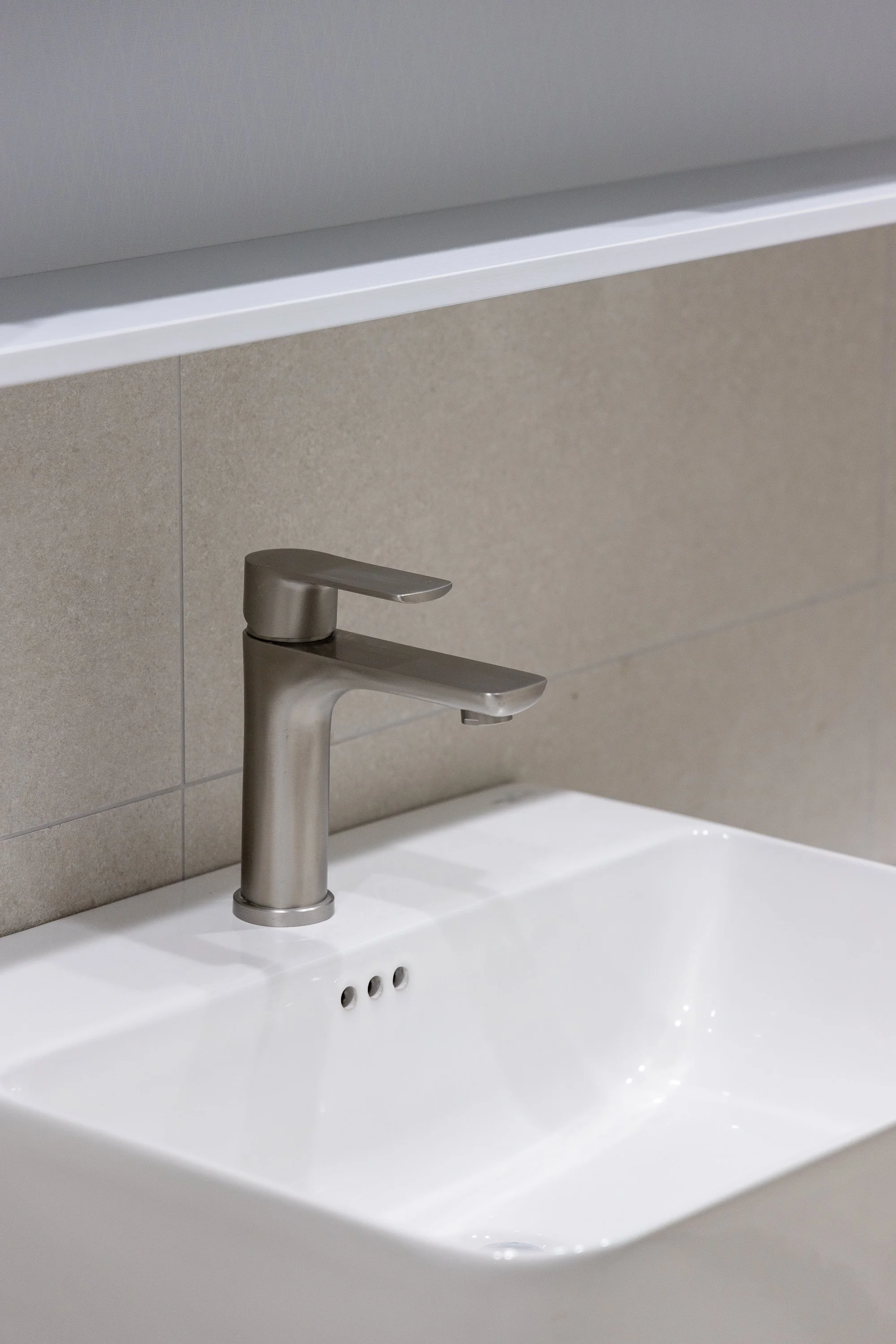 Close-up of a modern silver faucet mounted above a white bathroom sink with a beige tiled wall in the background.