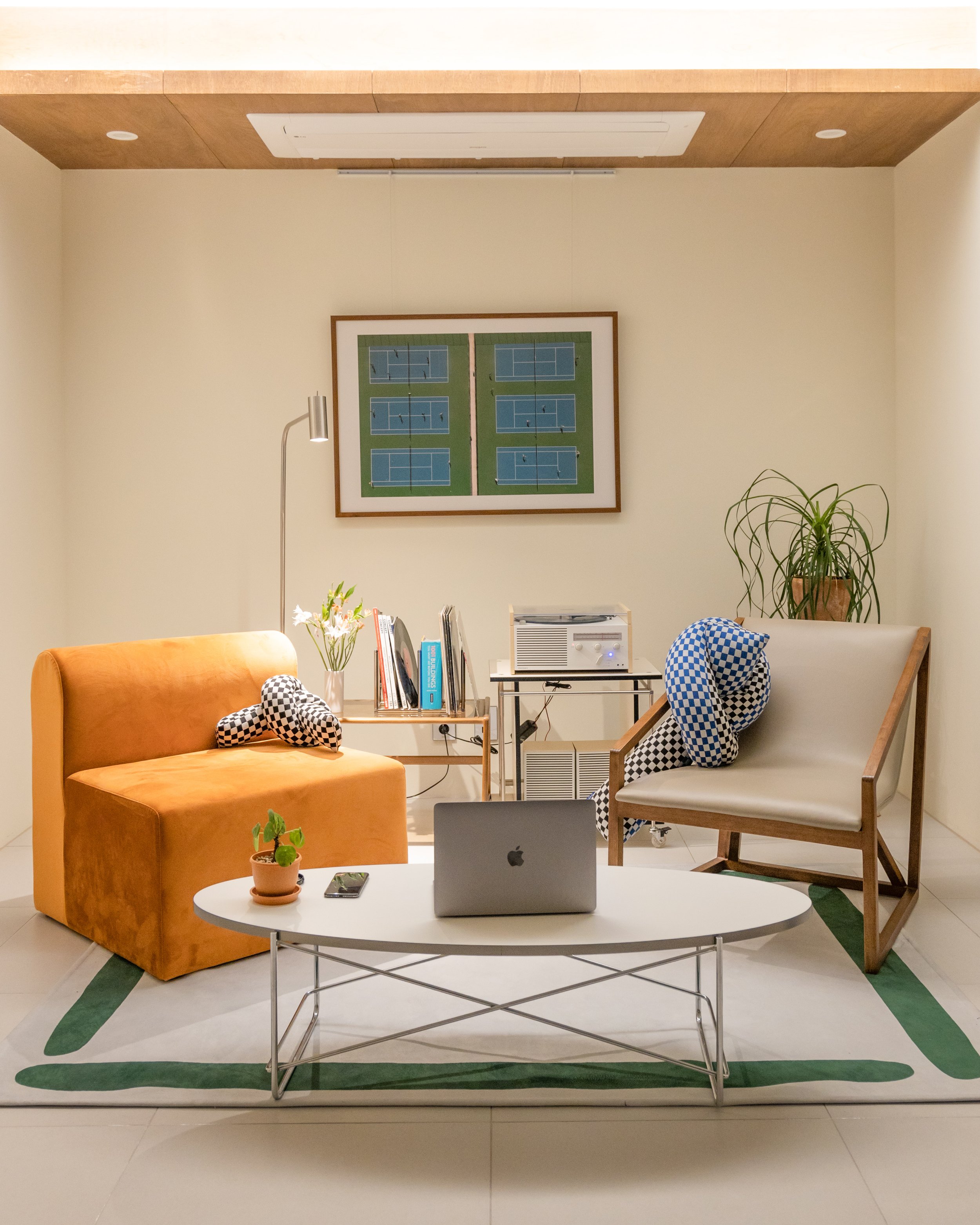 Living room with an orange velvet sofa, a white coffee table with a potted plant and laptop, two chairs with checkered cushions, a small side table with books and a record player, a large potted plant, and framed tennis court pictures on the wall.