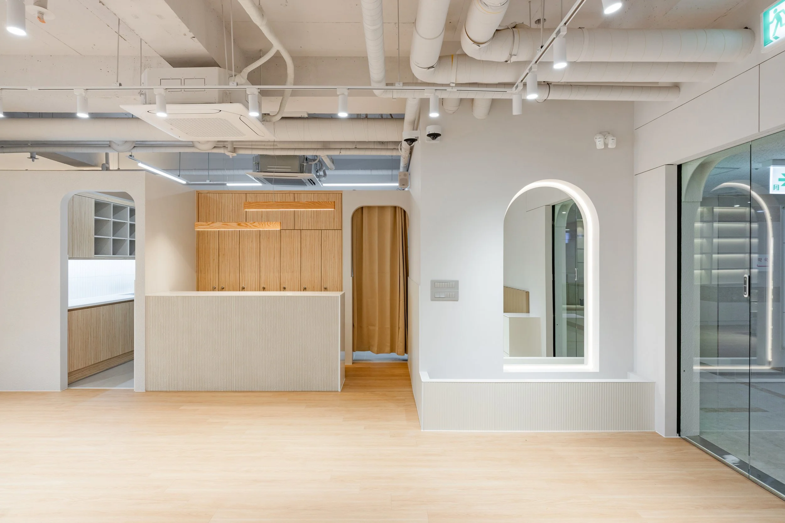 Modern interior space with wooden and white accents, featuring a reception area with a partial wall, built-in storage, arched reflective window, and glass sliding door, with exposed ceiling piping and bright lighting.