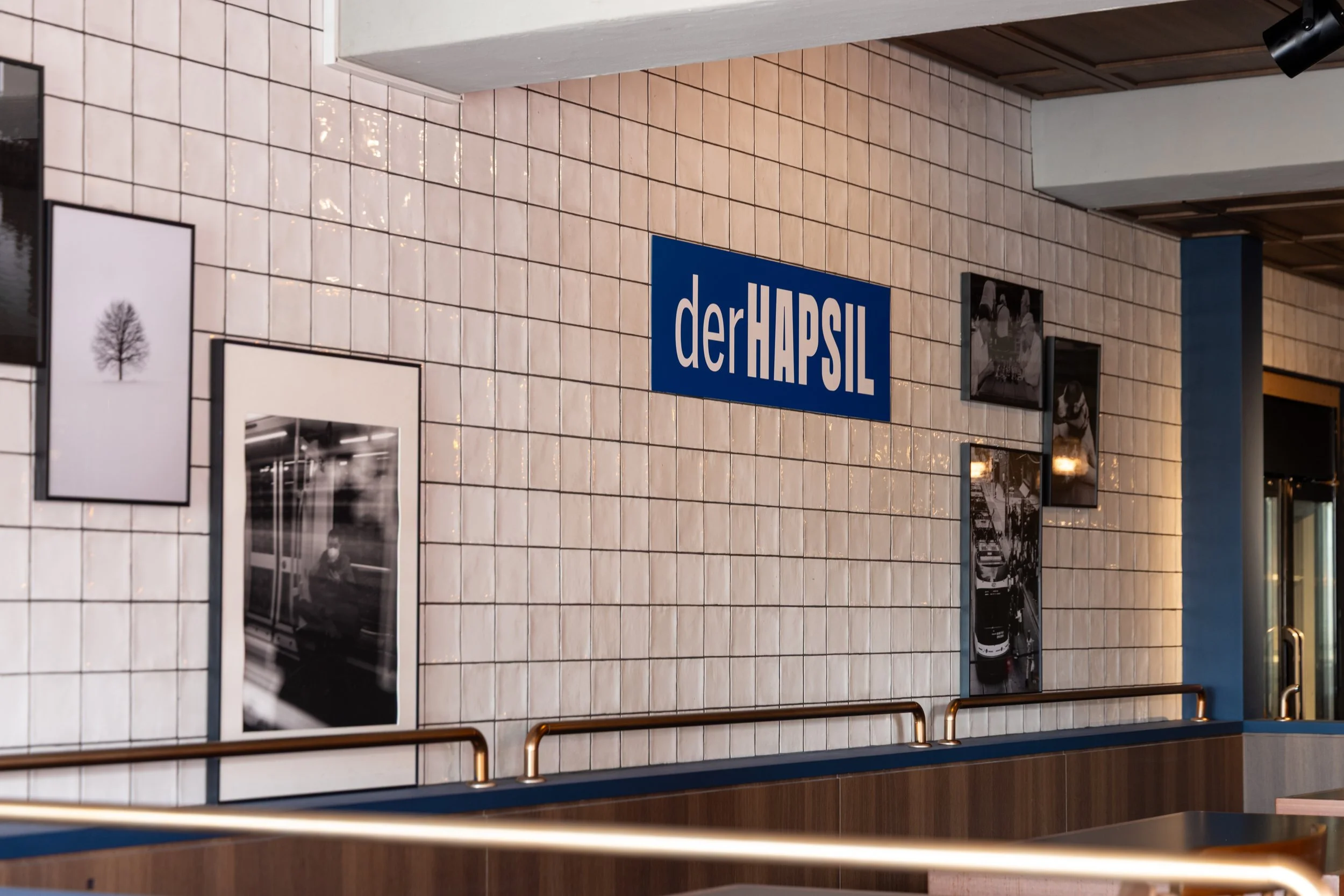 Interior of a building with a tiled wall, featuring black and white framed photos, a blue sign reading 'derHAPSIL', and a glass door with reflections.