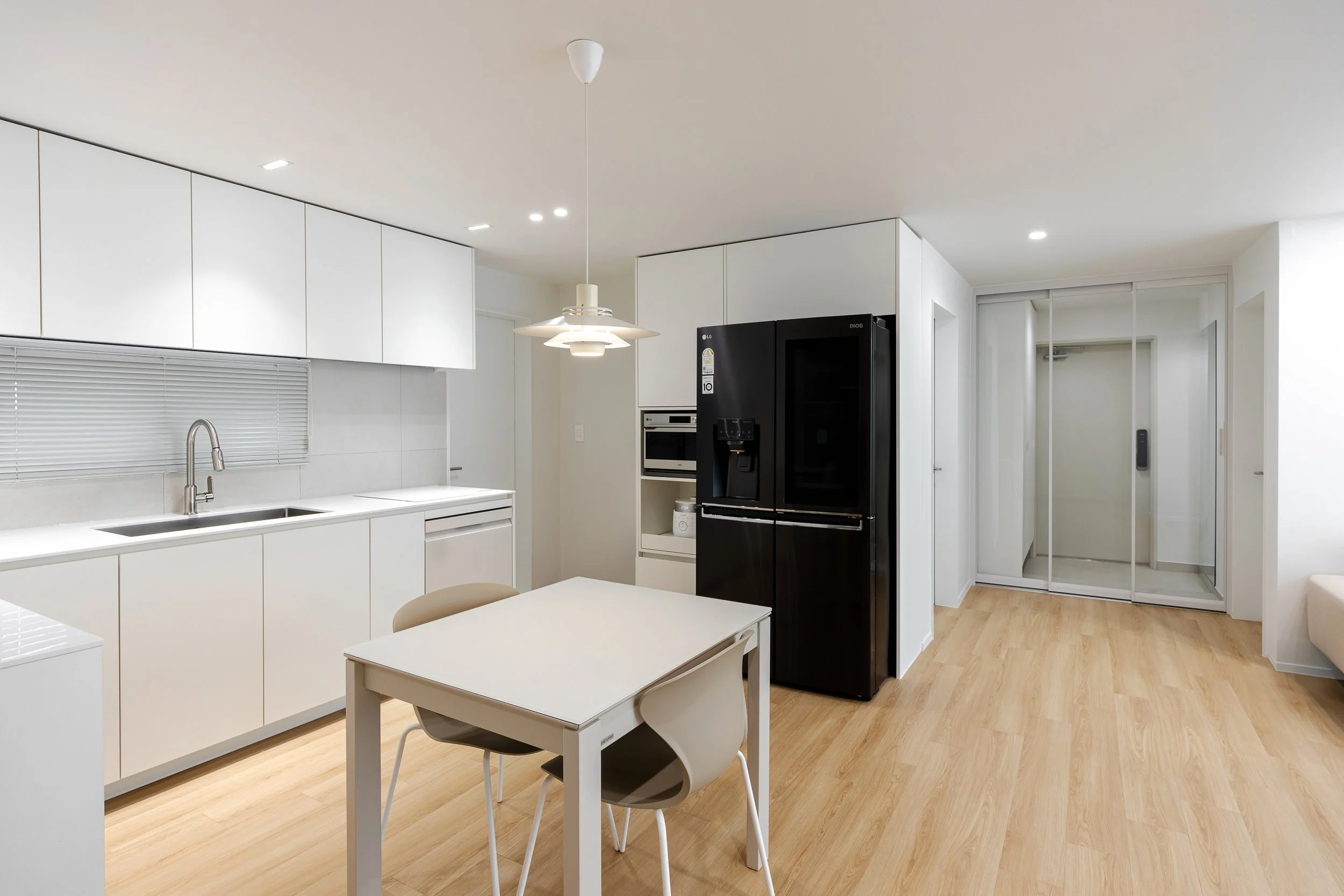 Modern kitchen with white cabinets, light wooden flooring, a black refrigerator, a small white dining table with two chairs, and a mirrored closet door.
