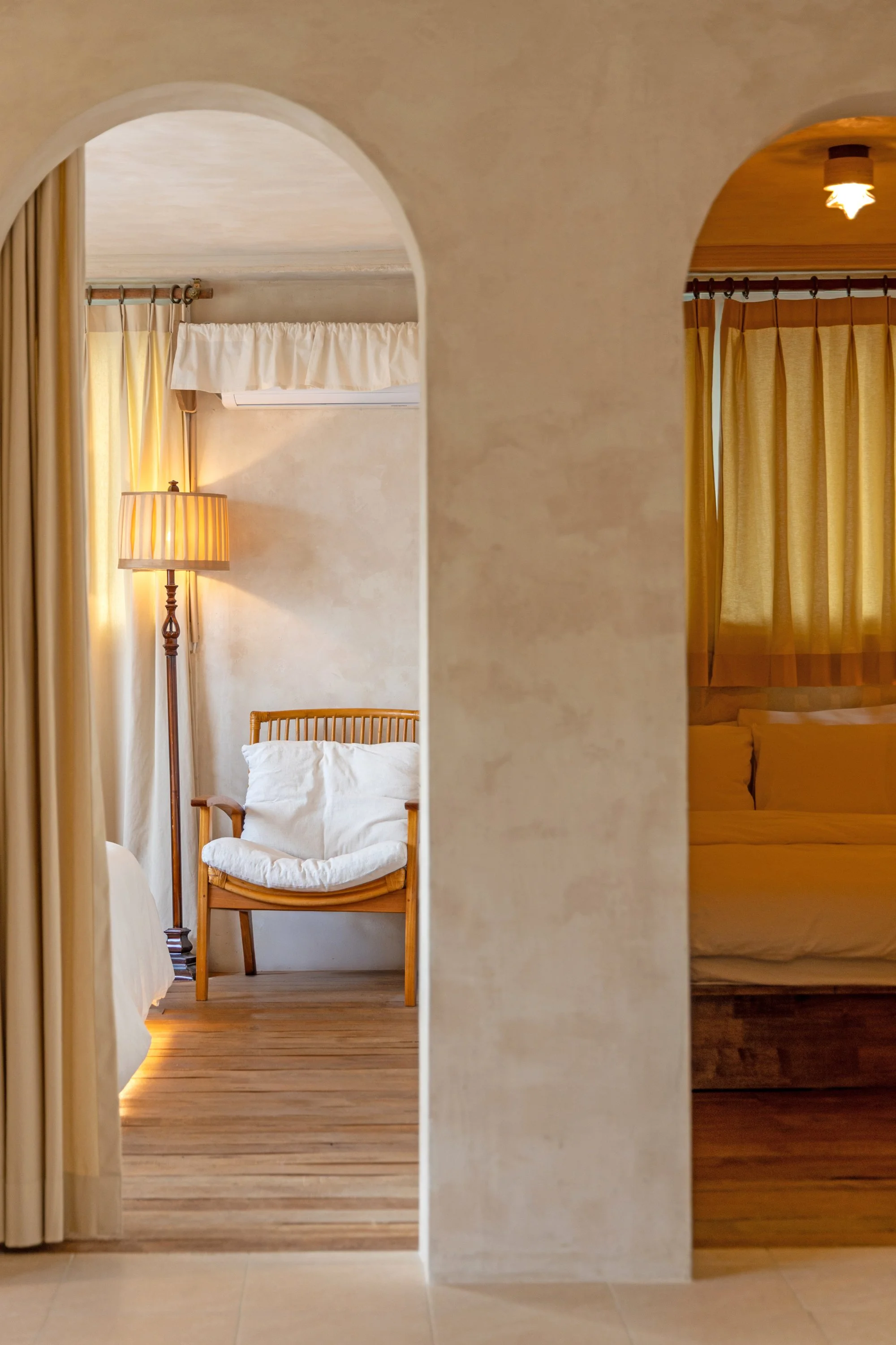A cozy interior view of a bedroom and sitting area separated by plastered archways, featuring a wooden chair with cushions, a standing lamp, and bed with curtains.