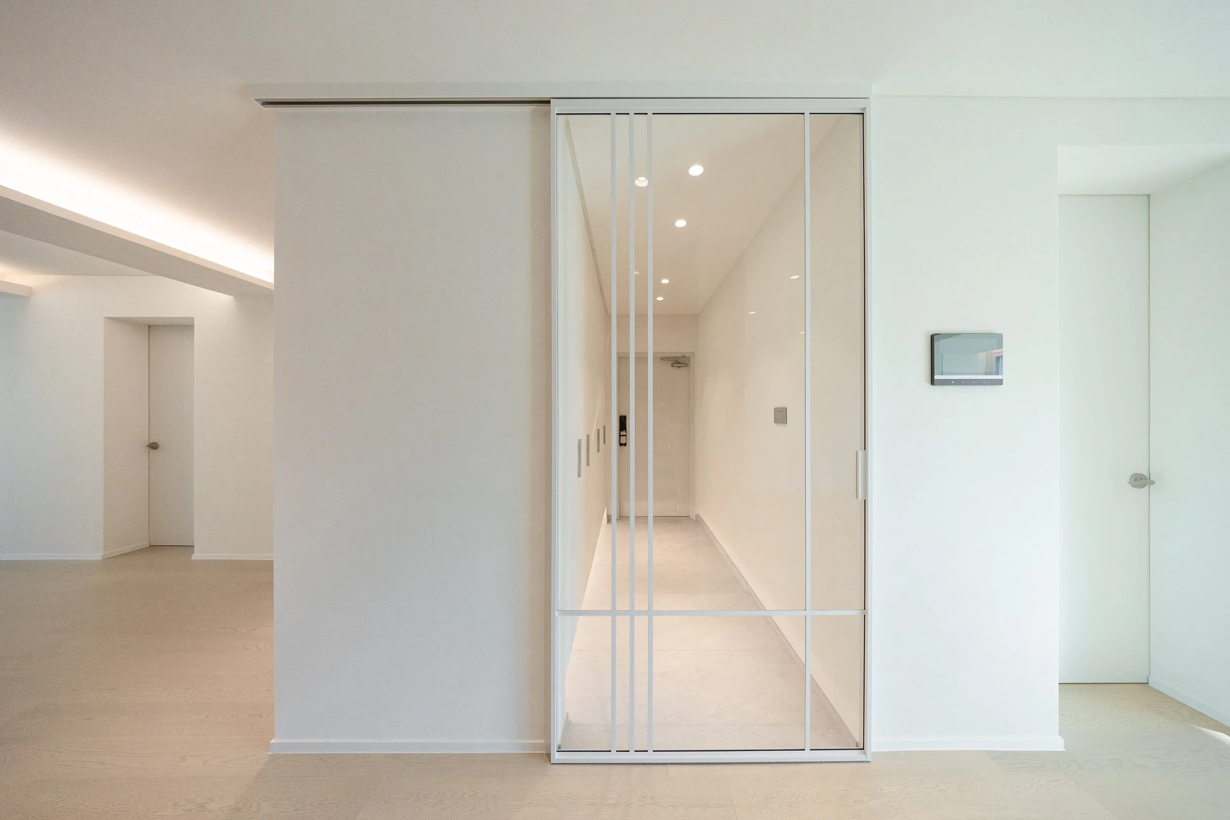 Modern minimalist entryway with white walls, light wood flooring, a white sliding door with glass panels and metal frame, and recessed ceiling lights.