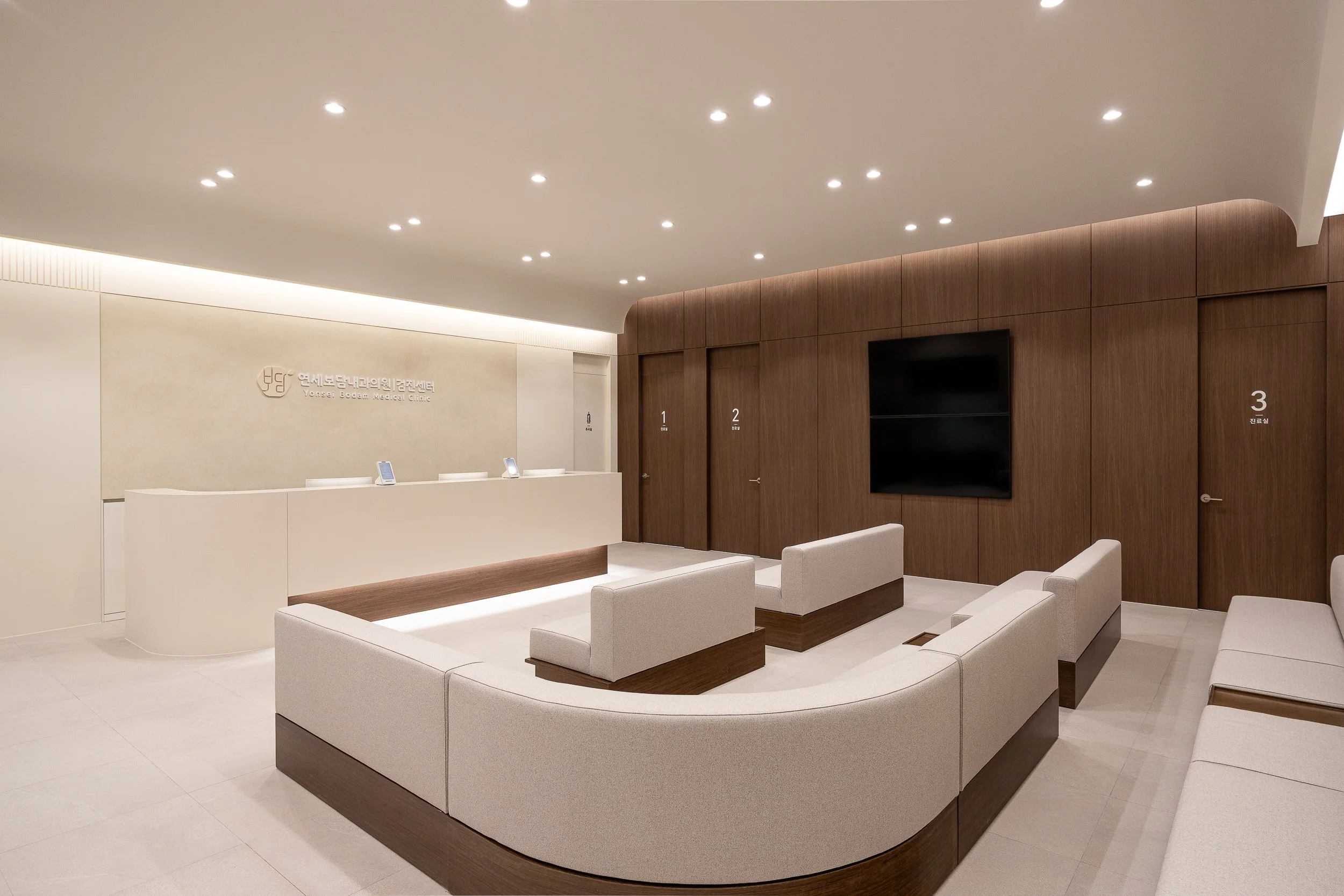 Modern medical clinic reception area with white and wood accents, waiting area with beige sofas, and a black wall-mounted TV.