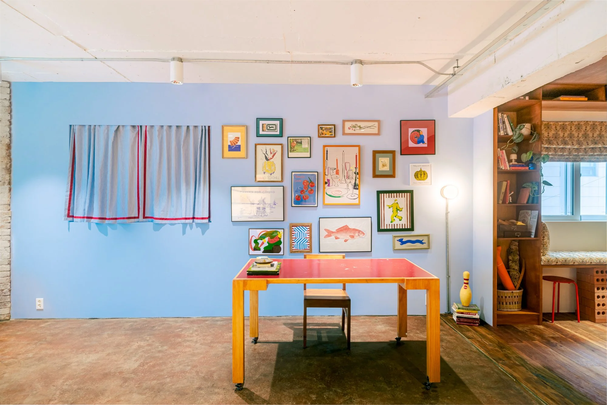 A room with a blue wall decorated with various framed animal and art prints, a small window with a curtain, a red table with a chair, a bookshelf with plants and books, and a vintage bowling pin lamp.