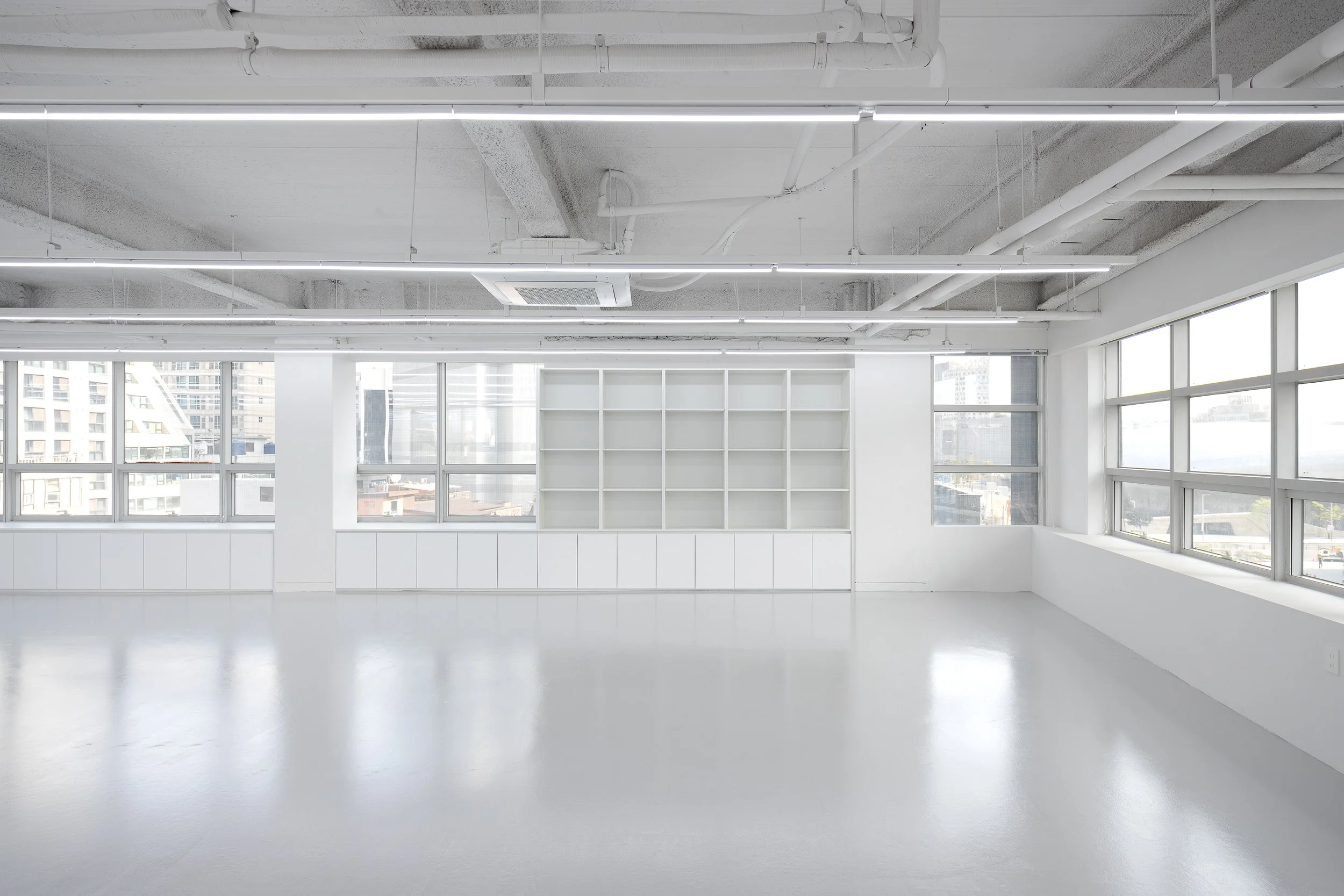 Empty modern office room with white walls, large windows, white shelving unit, and ceiling with exposed pipes and lighting.