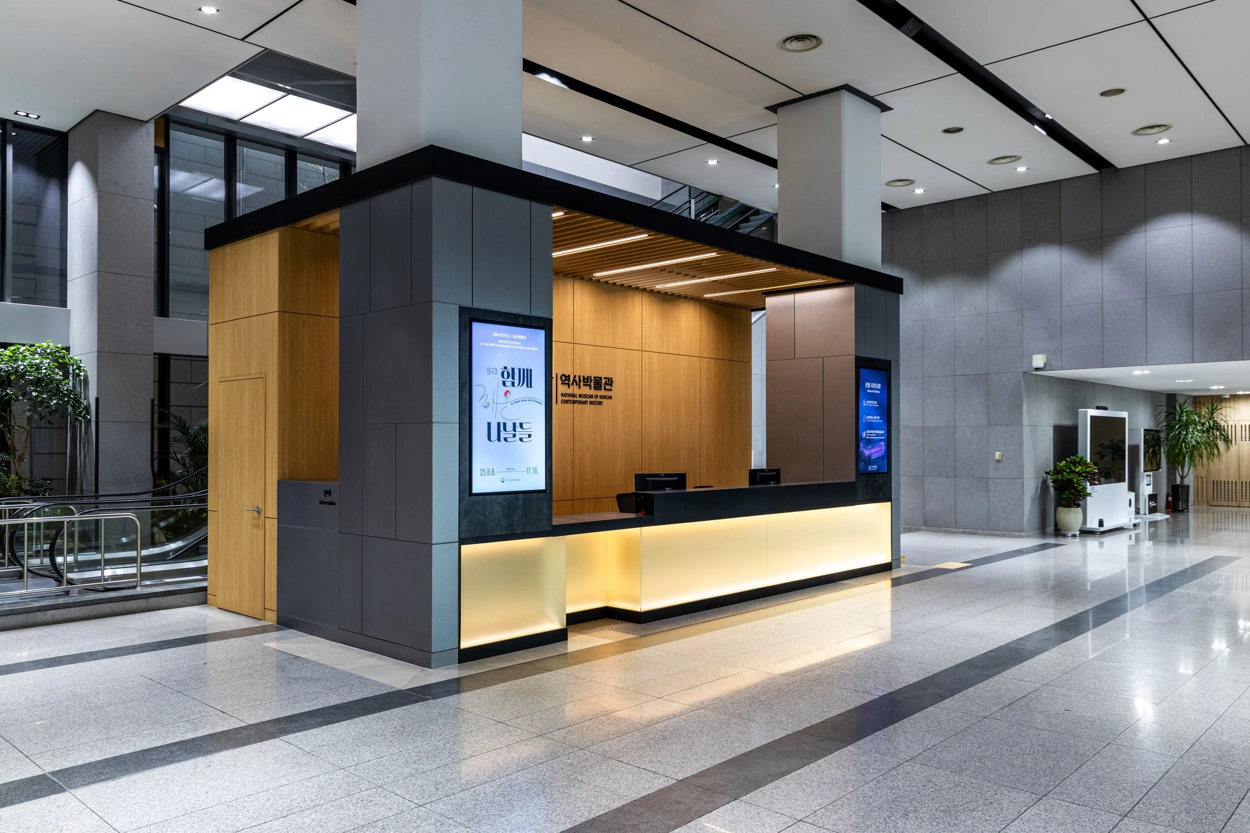 Reception desk in a modern building with gray walls, wooden accents, and digital screens displaying information.