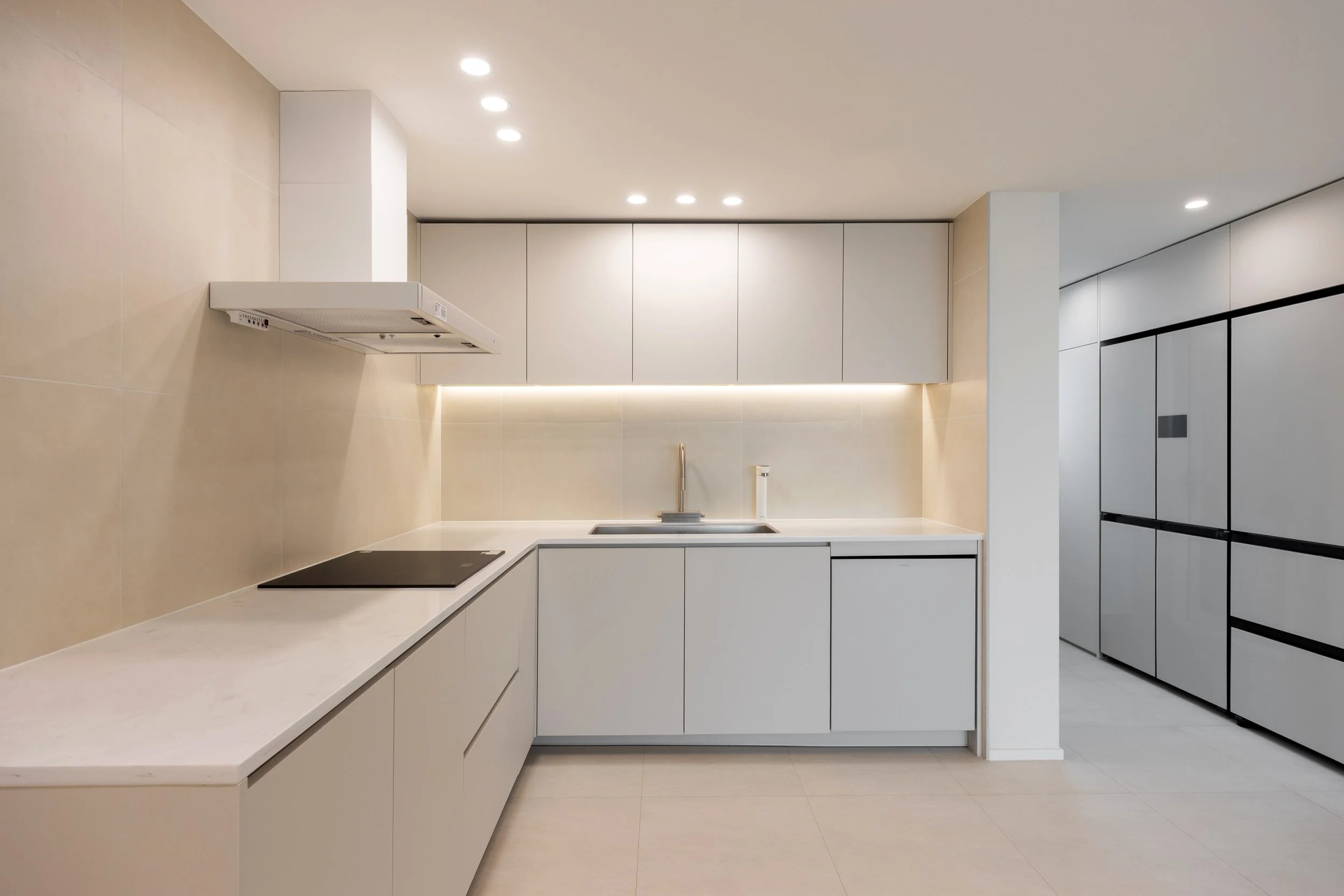 Modern minimalist kitchen with white cabinets, beige backsplash, black stovetop, and built-in refrigerators on the right, illuminated by ceiling and under-cabinet lighting.