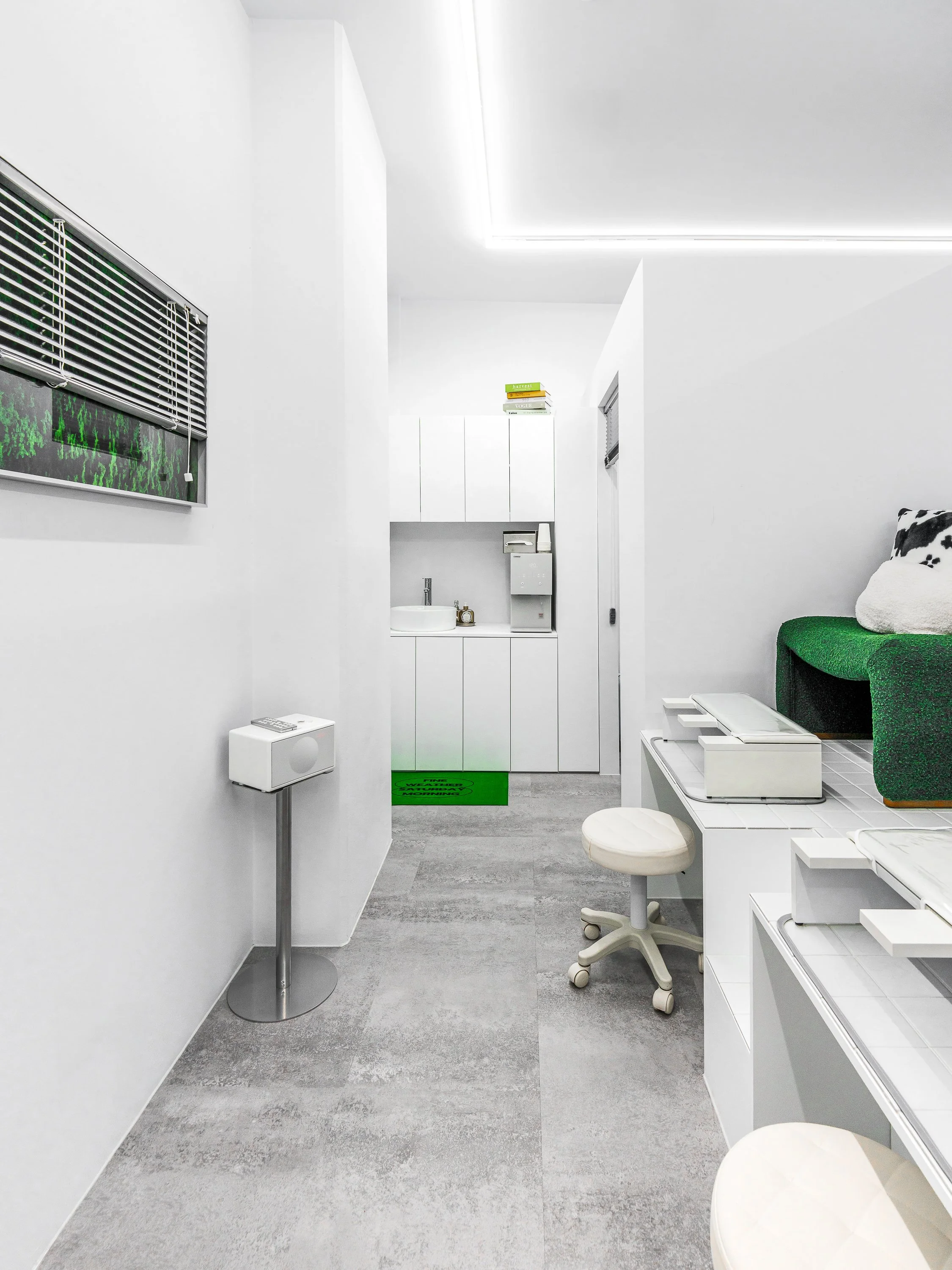 Minimalist medical or office space with white walls, a window with a metal grill, a small white side table with a remote control, a white cabinet with a sink, a green mat, a white chair, and a green couch with a cow-patterned pillow.