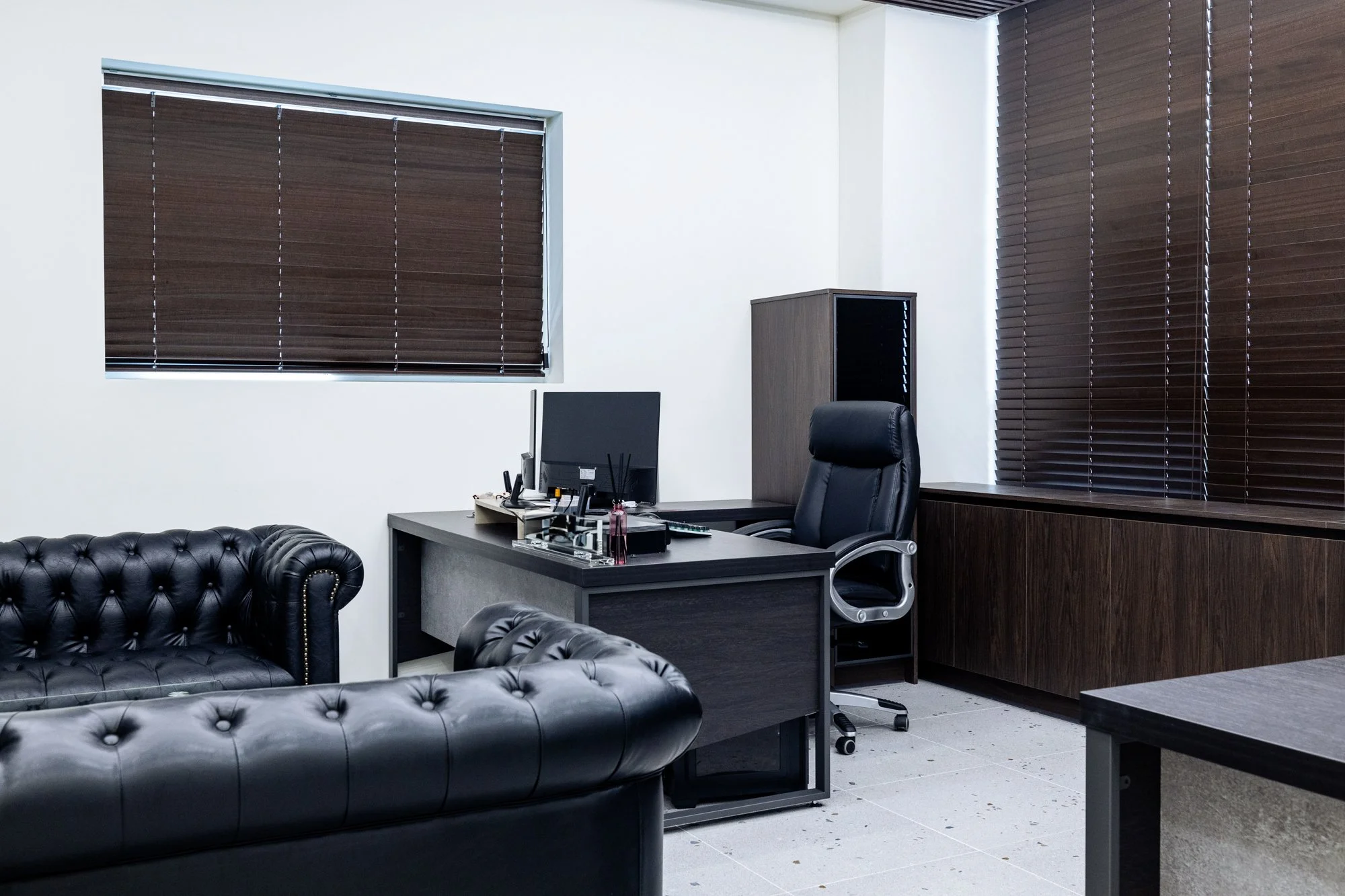 An office with dark wood blinds, a black leather couch, a desk with a computer, and a black office chair.
