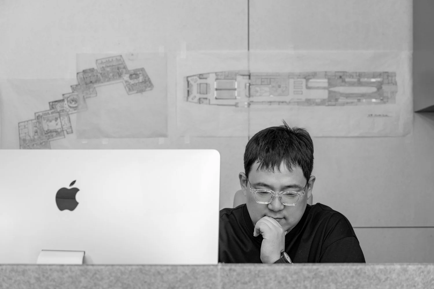 A person with short dark hair, wearing glasses and a black shirt, sitting at a desk with an Apple computer. Behind them are architectural drawings or blueprints taped to the wall.