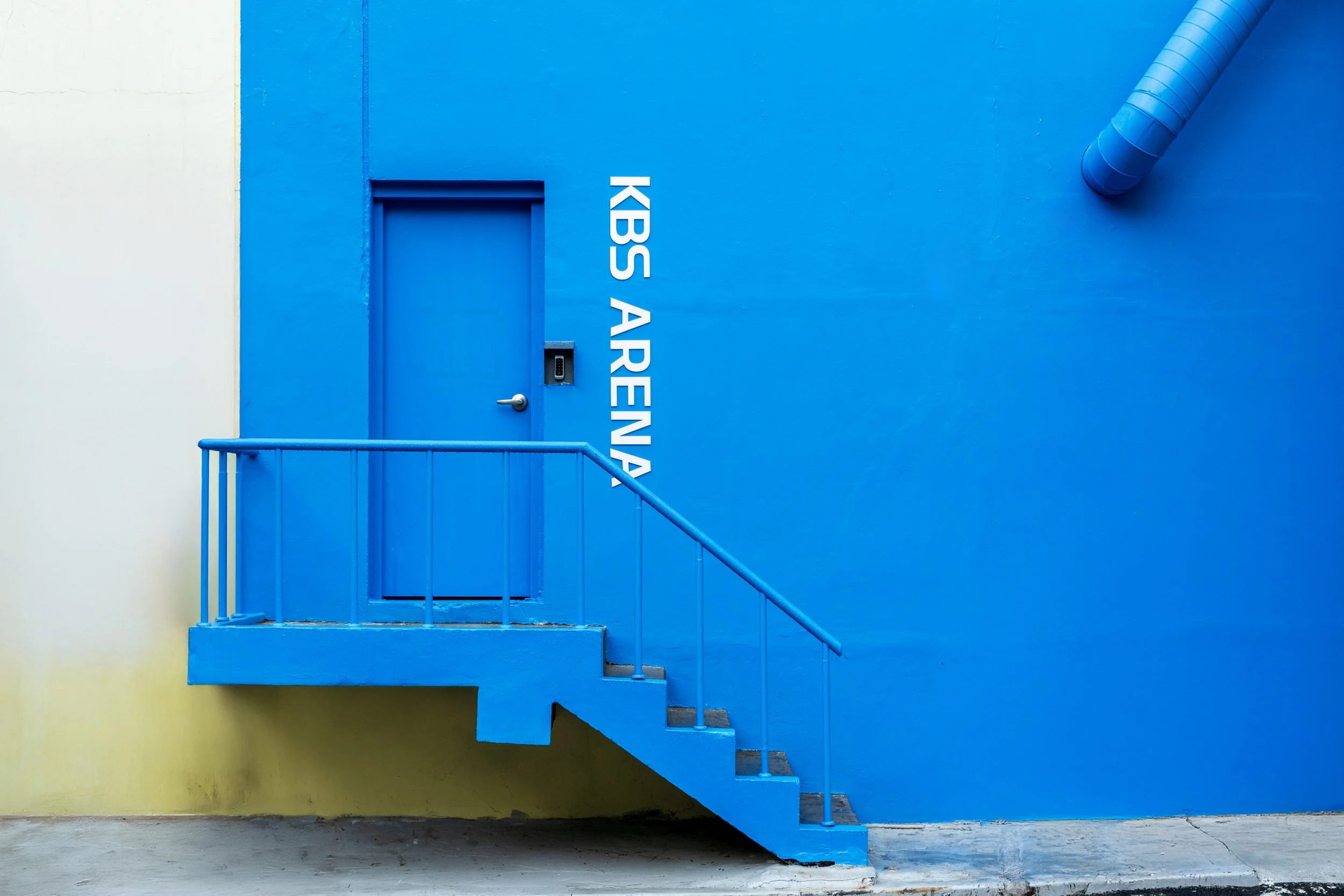 Blue building with stairs, door, and signage reading 'KBS ARENA' in white letters, with a blue ventilation duct on the upper right.