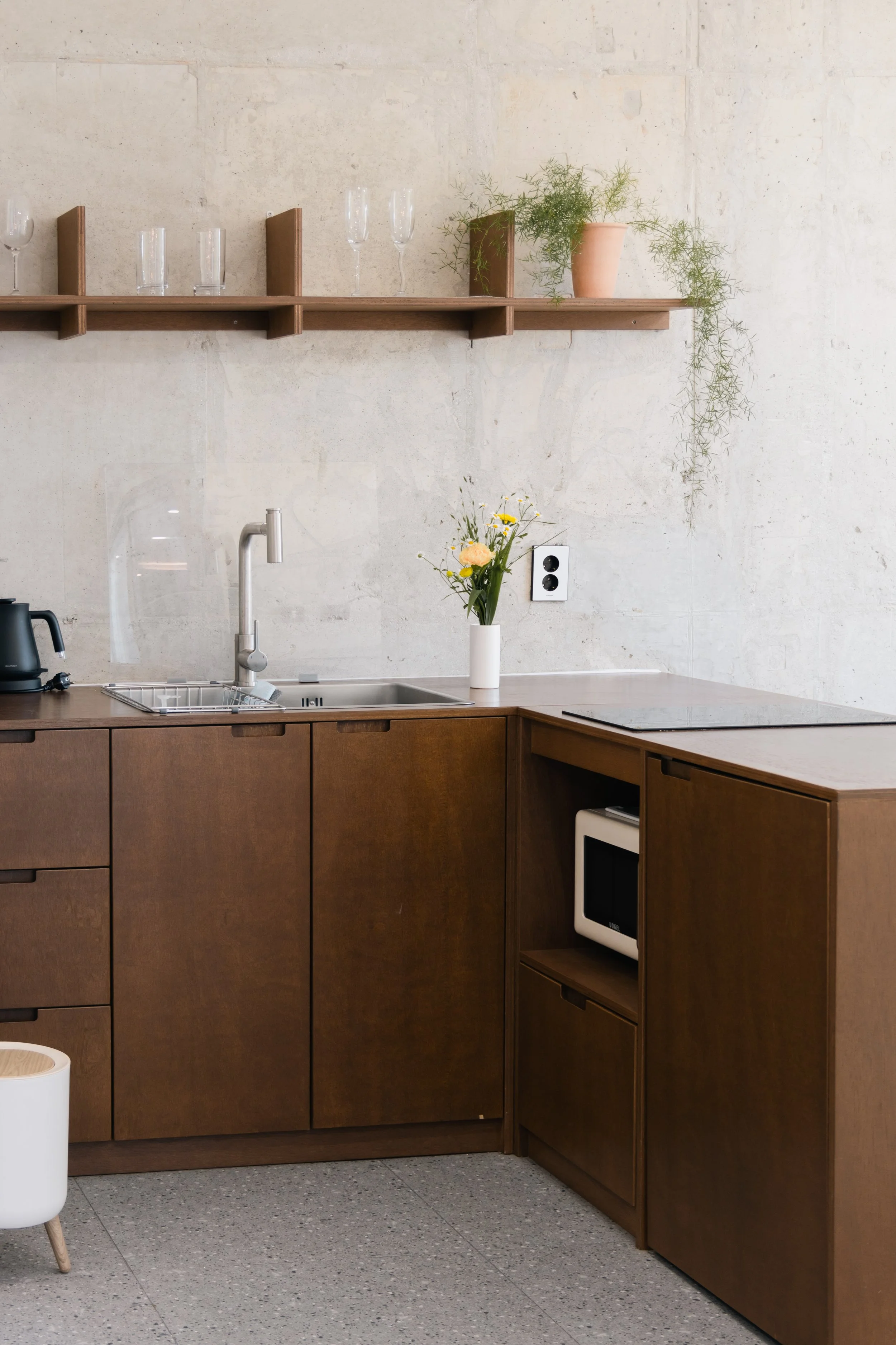 Minimalist kitchen with dark wood cabinets, a vase of flowers, a stovetop, microwave, and a shelf with glasses and a plant.