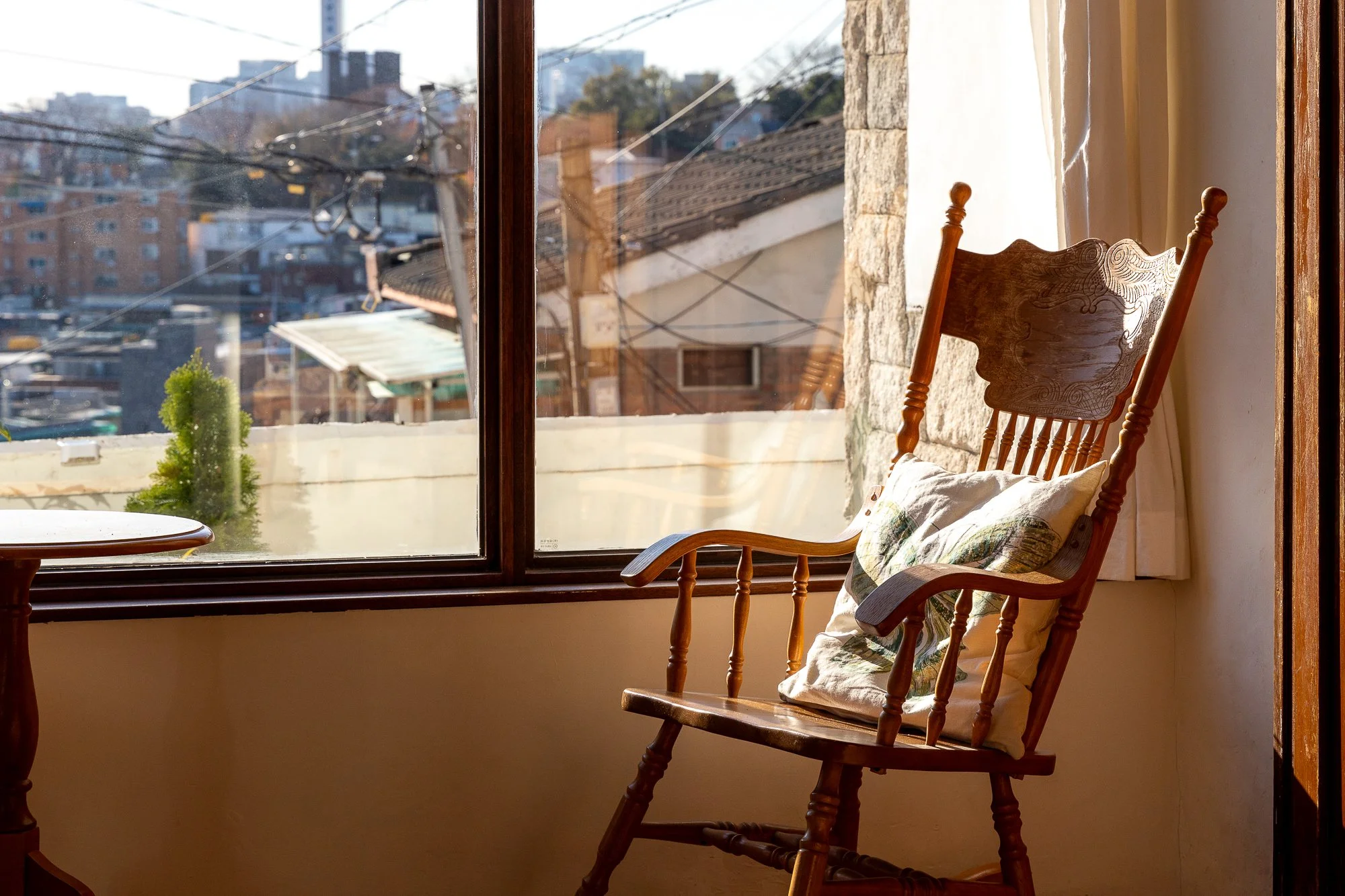 A wooden rocking chair with a floral pillow sits next to a large window in a room with stone and white walls. Outside the window, a view of neighboring houses, power lines, and a cityscape is visible.