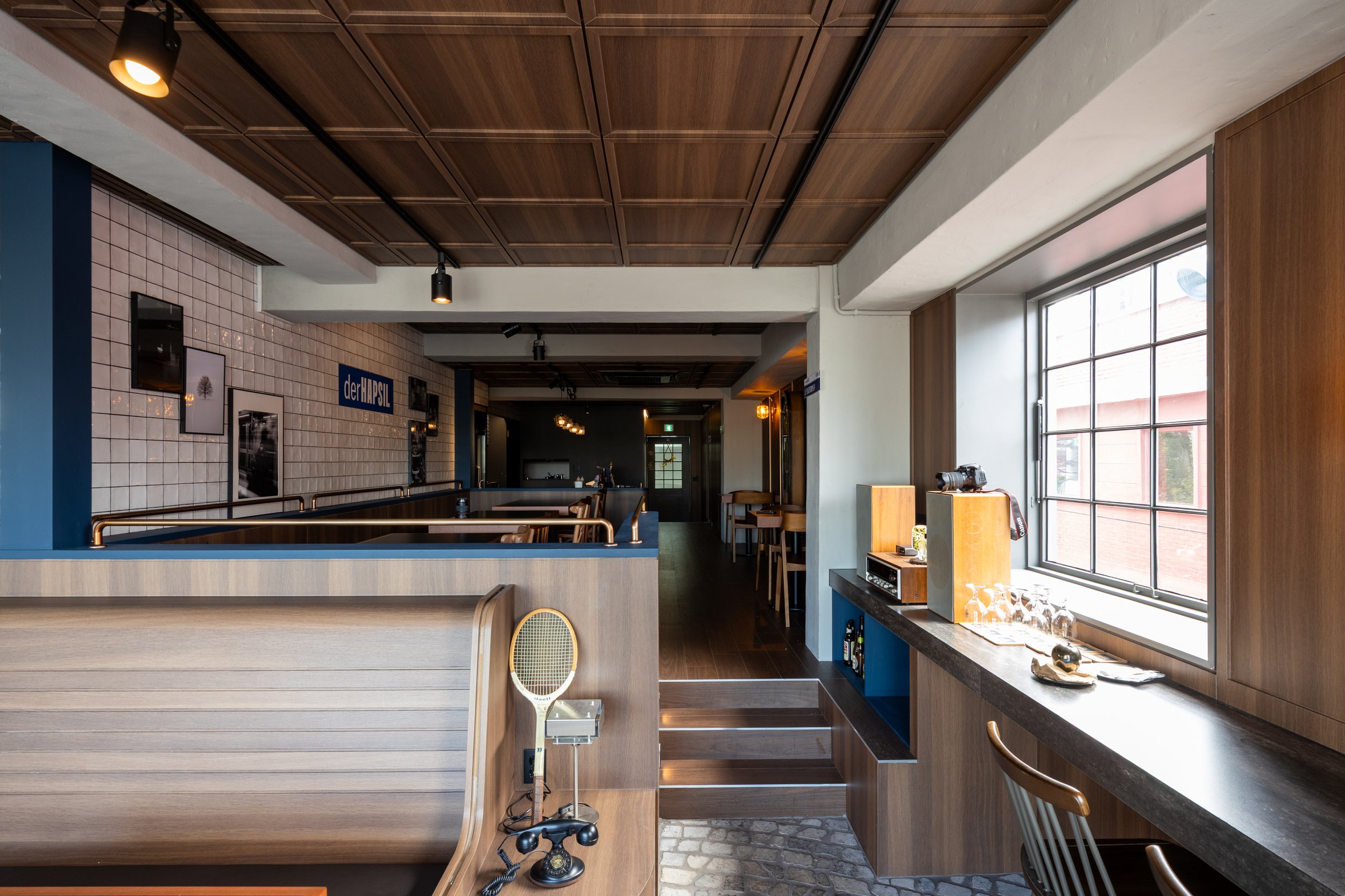 Interior of a modern, cozy café or restaurant with wooden accents, a large window, and various seating options. There are wall decorations, a TV, bar stools, and a counter with glasses. The space has warm lighting and a stylish, inviting atmosphere.