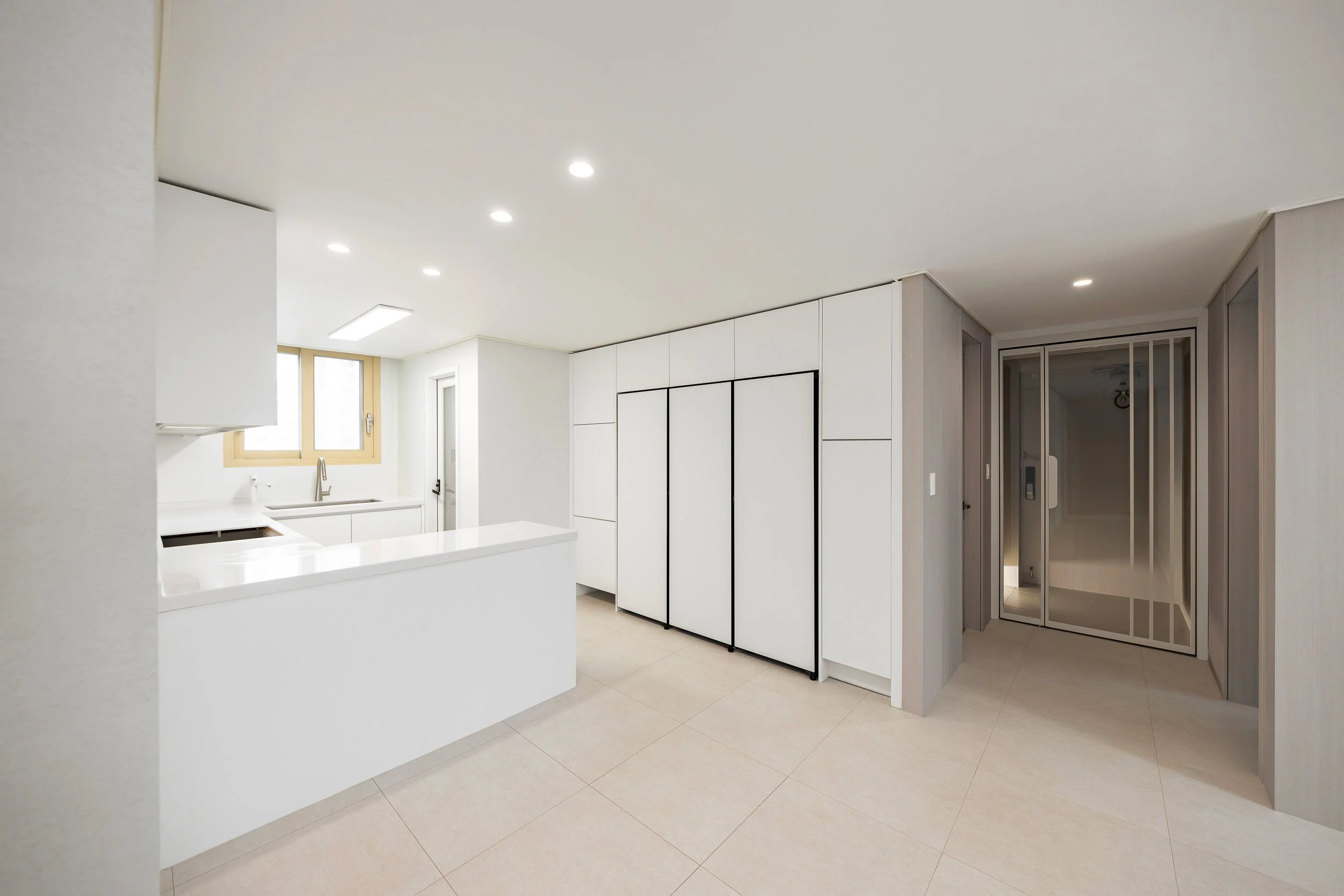 Minimalist kitchen with white cabinets, a window above the sink, and an open counter, adjacent to a hallway with a glass door.
