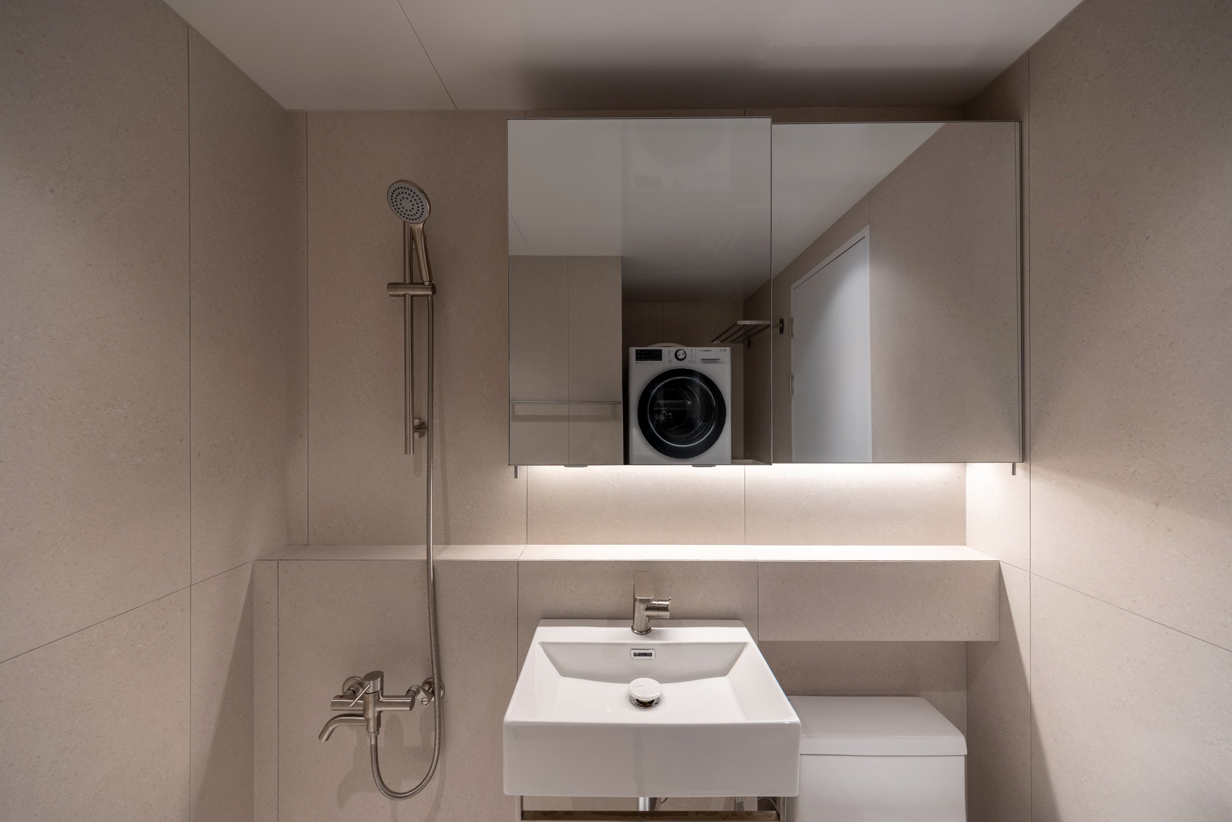 A minimalist bathroom with beige tiled walls and a large mirror. Includes a white rectangular sink, a stainless steel faucet, and a washing machine reflected in the mirror.