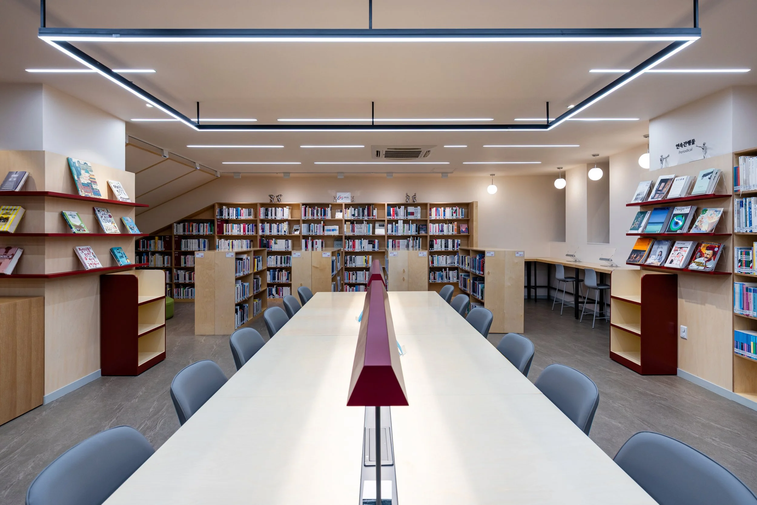 Interior of a modern library with bookshelves, a long central table with chairs, and a reading area with a bar-style counter and stools.