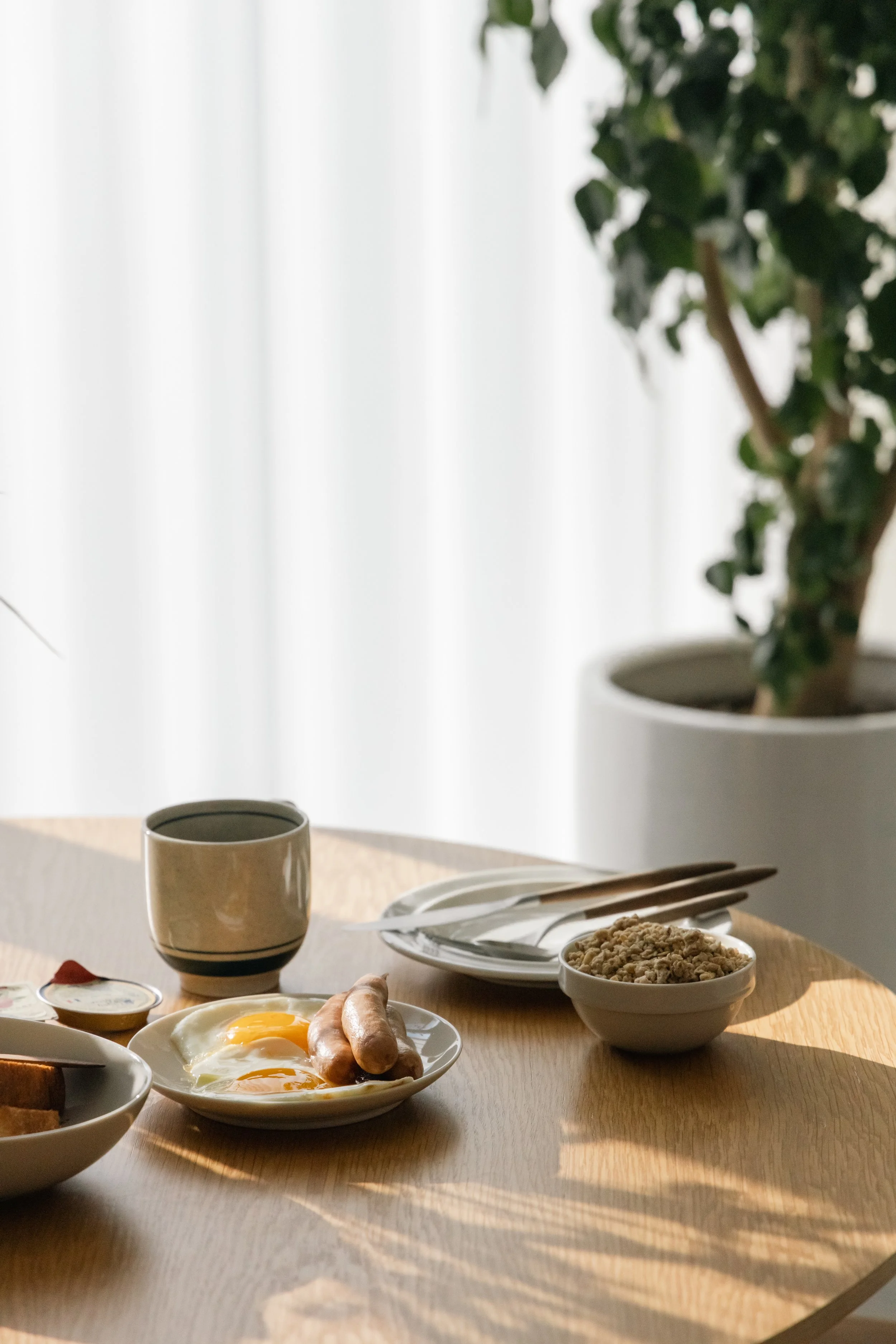 Breakfast table with eggs, sausages, cereal, toast, a cup, and cutlery in front of a potted plant by a window with sheer curtains.