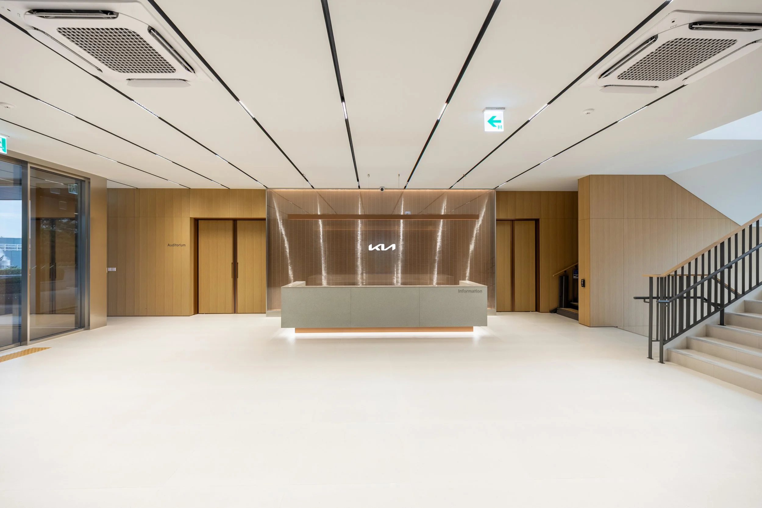 Modern interior of a Kia dealership lobby with a reception desk, wooden wall panels, and glass doors leading to an auditorium and other rooms. Staircase with black railing on the right.