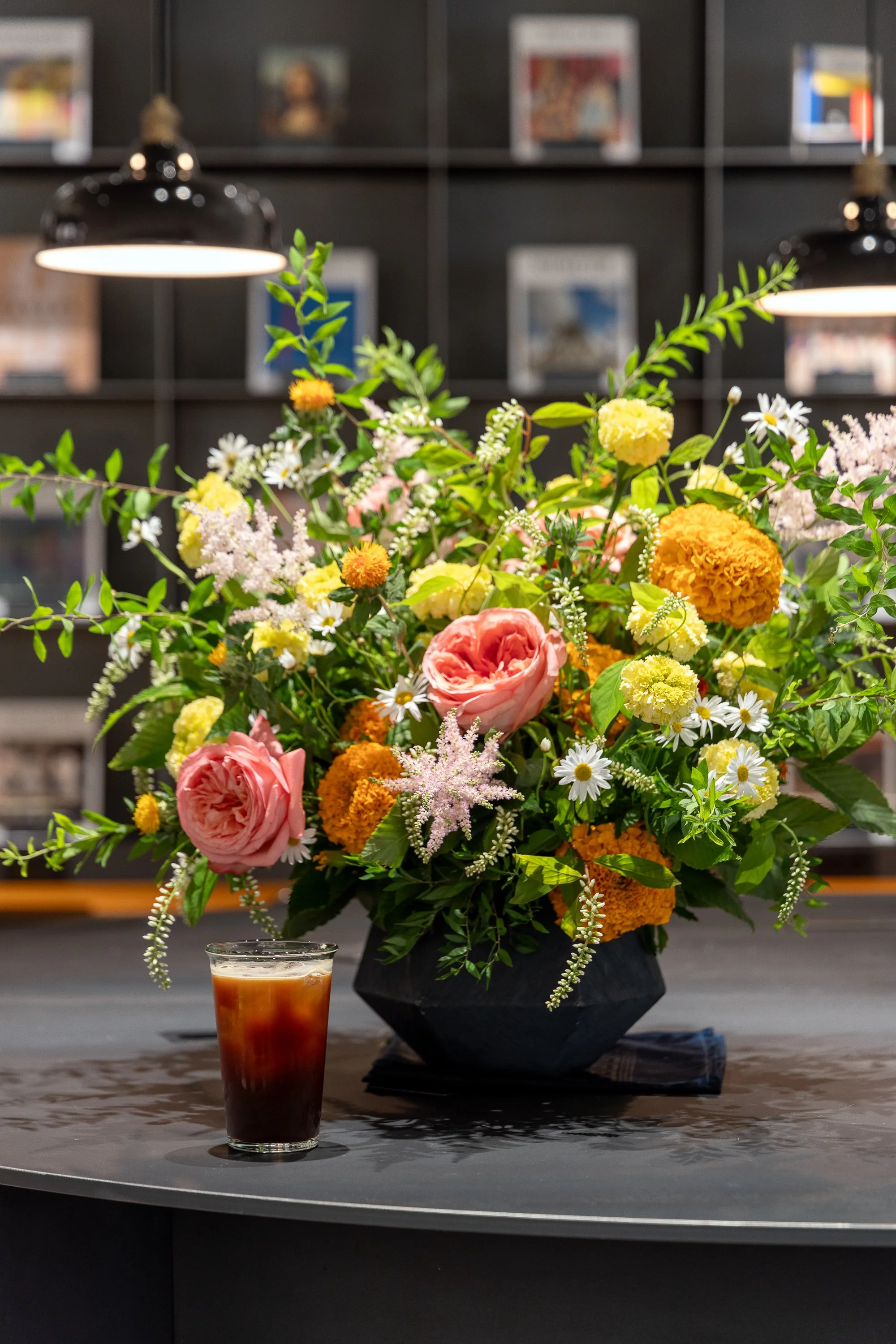 A large bouquet of various colorful flowers, including pink roses, orange marigolds, and white daisies, arranged in a black vase on a black table, with a glass of iced coffee in the foreground and a dark bookshelf and hanging lights in the background