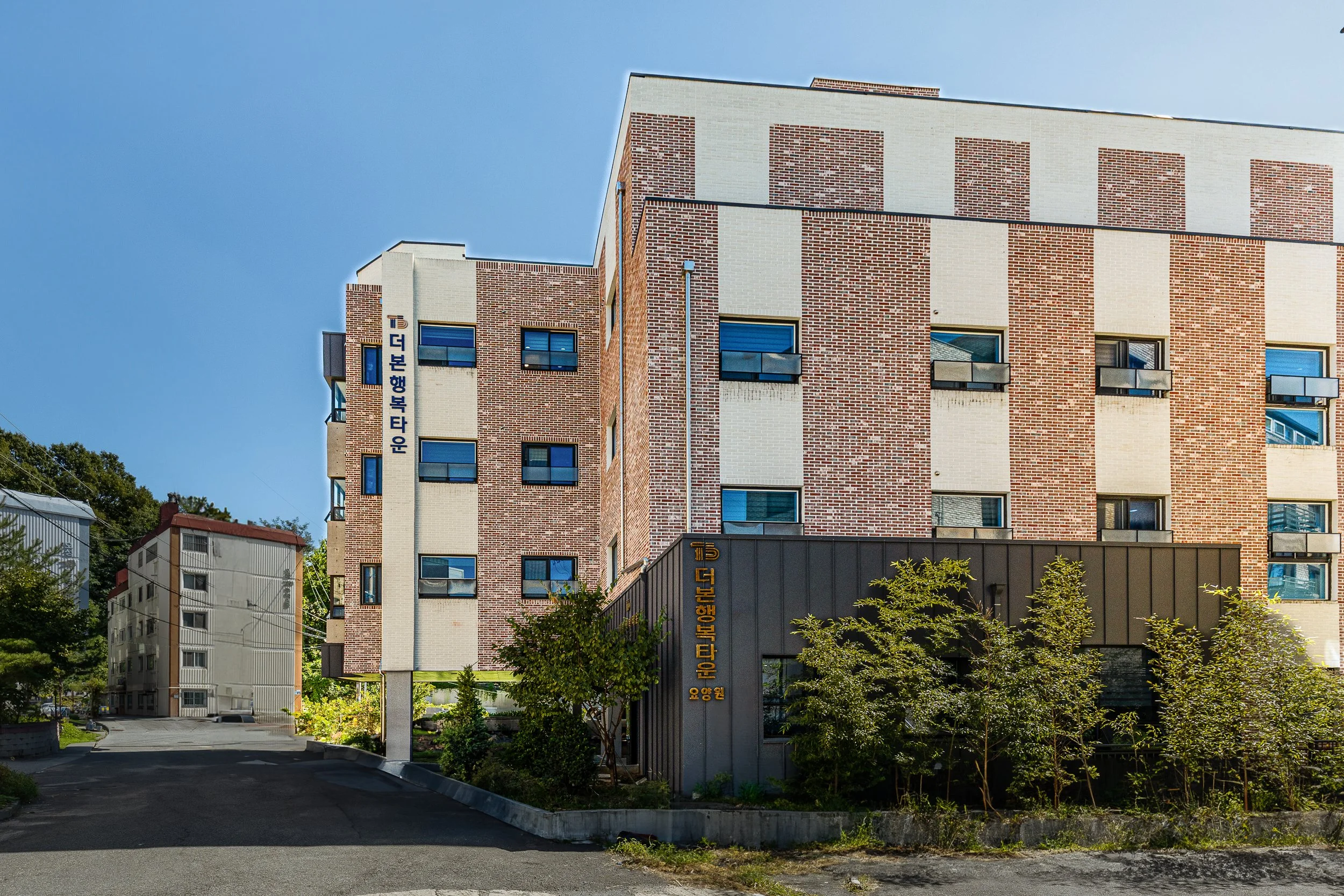 A modern multi-story building with brick and beige exterior, surrounded by trees and bushes, under a clear blue sky.