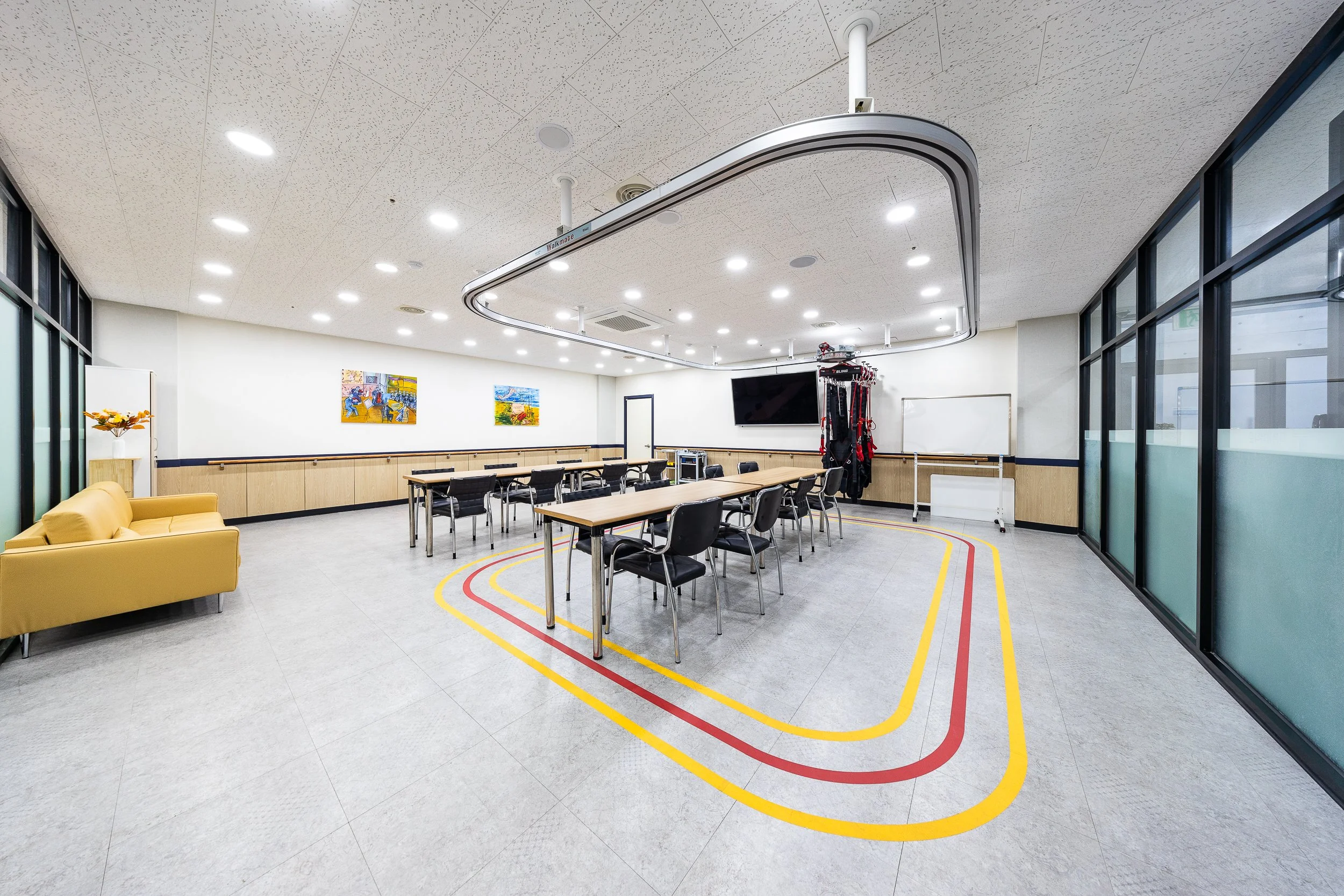 A spacious meeting room with tables and chairs arranged in a U-shape, a yellow sofa on the left, a wall-mounted TV, colorful artwork on the walls, large glass windows on the right, and a circular ceiling fixture with lighting.