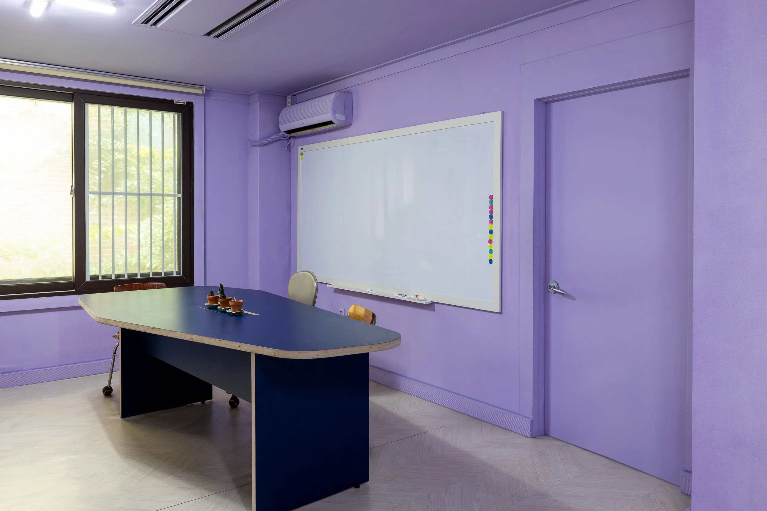 Empty classroom with purple walls, window with bars, whiteboard, desk with three potted cacti, and an air conditioning unit.