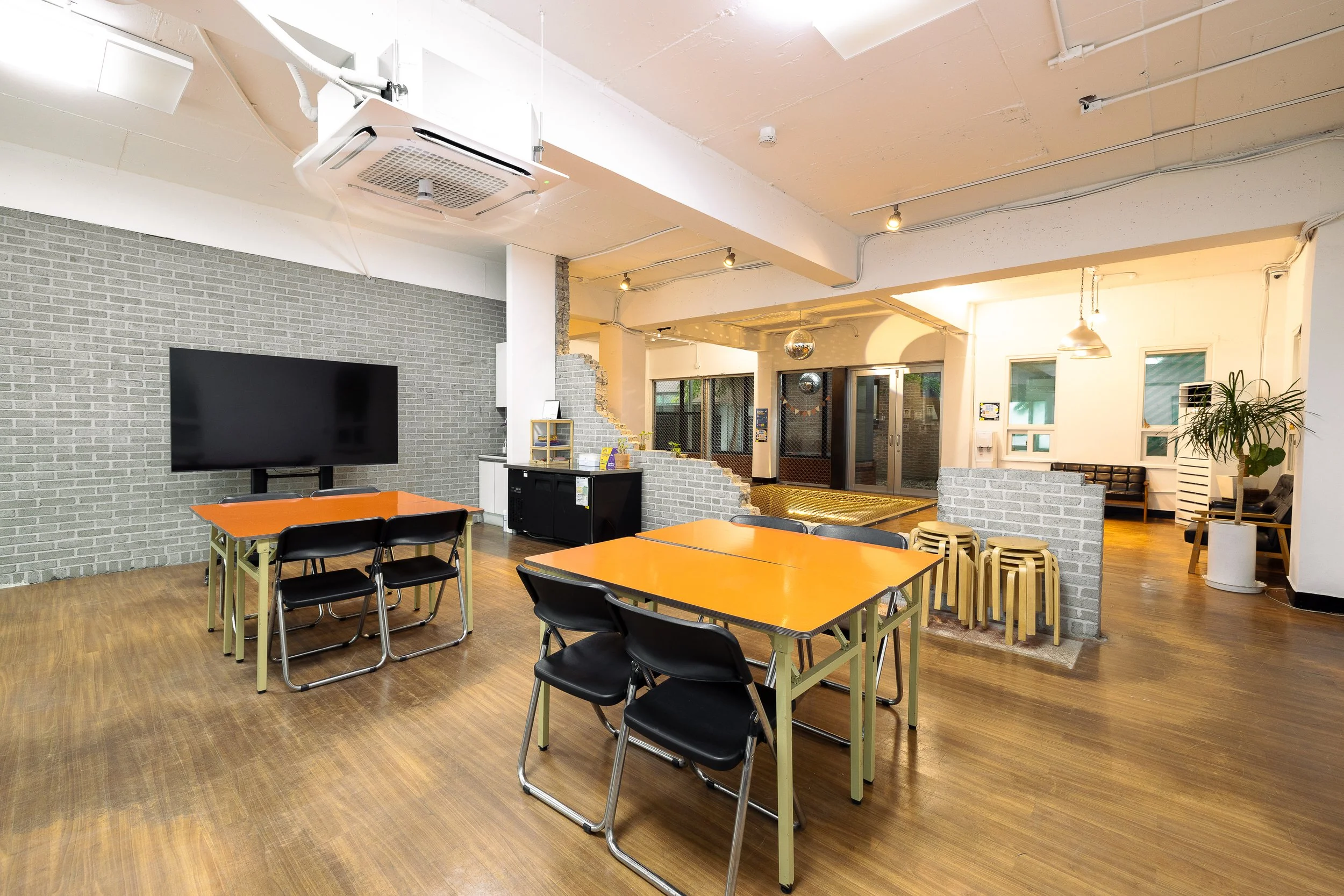 Interior of a modern lounge or meeting room with wooden flooring, orange tables, black chairs, a large wall-mounted TV, a server station, small stacked stools, and potted plants, illuminated by ceiling lights and pendant lamps.