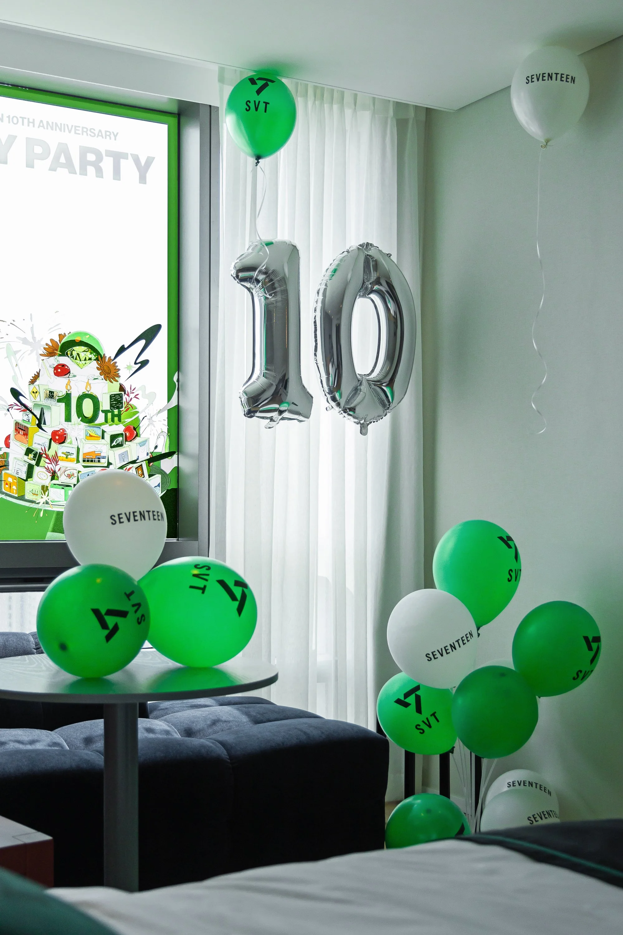 Celebration decorations for the 10th anniversary of the K-pop group Seventeen, including large silver balloons forming the number 10, and green and white balloons with the group's logo and name, set in a room with a window and curtains.